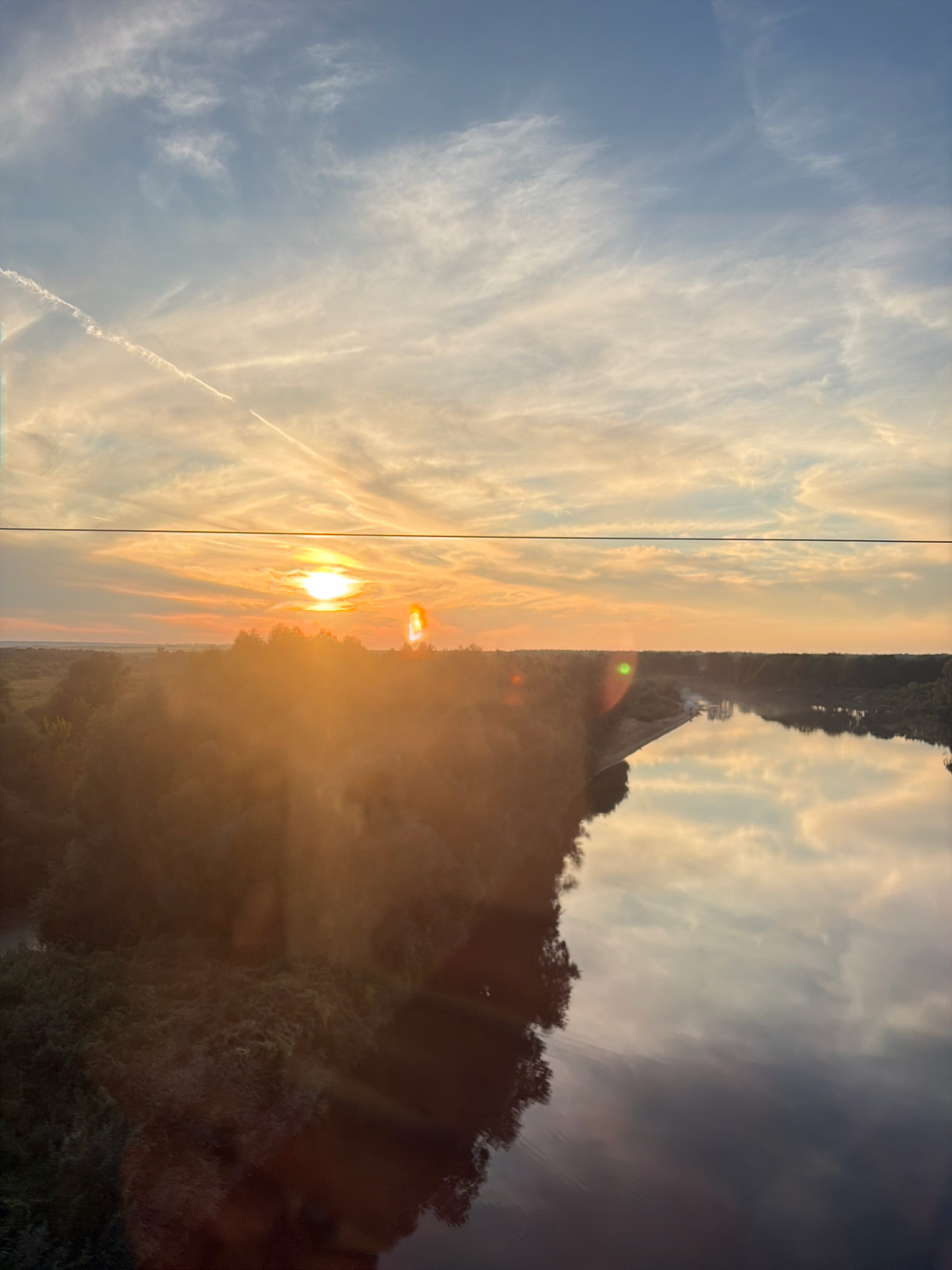 The image depicts a wide view of the Potiomkin River in Solovyovo, Russia, at sunset. The sky is a gradient of blue and orange, with wispy clouds and contrails from an aircraft. The sun is low on the horizon, casting a warm glow over the landscape. Trees line the riverbanks, their reflections visible in the calm water. The overall scene is tranquil and picturesque, conveying the beauty of nature at dusk.