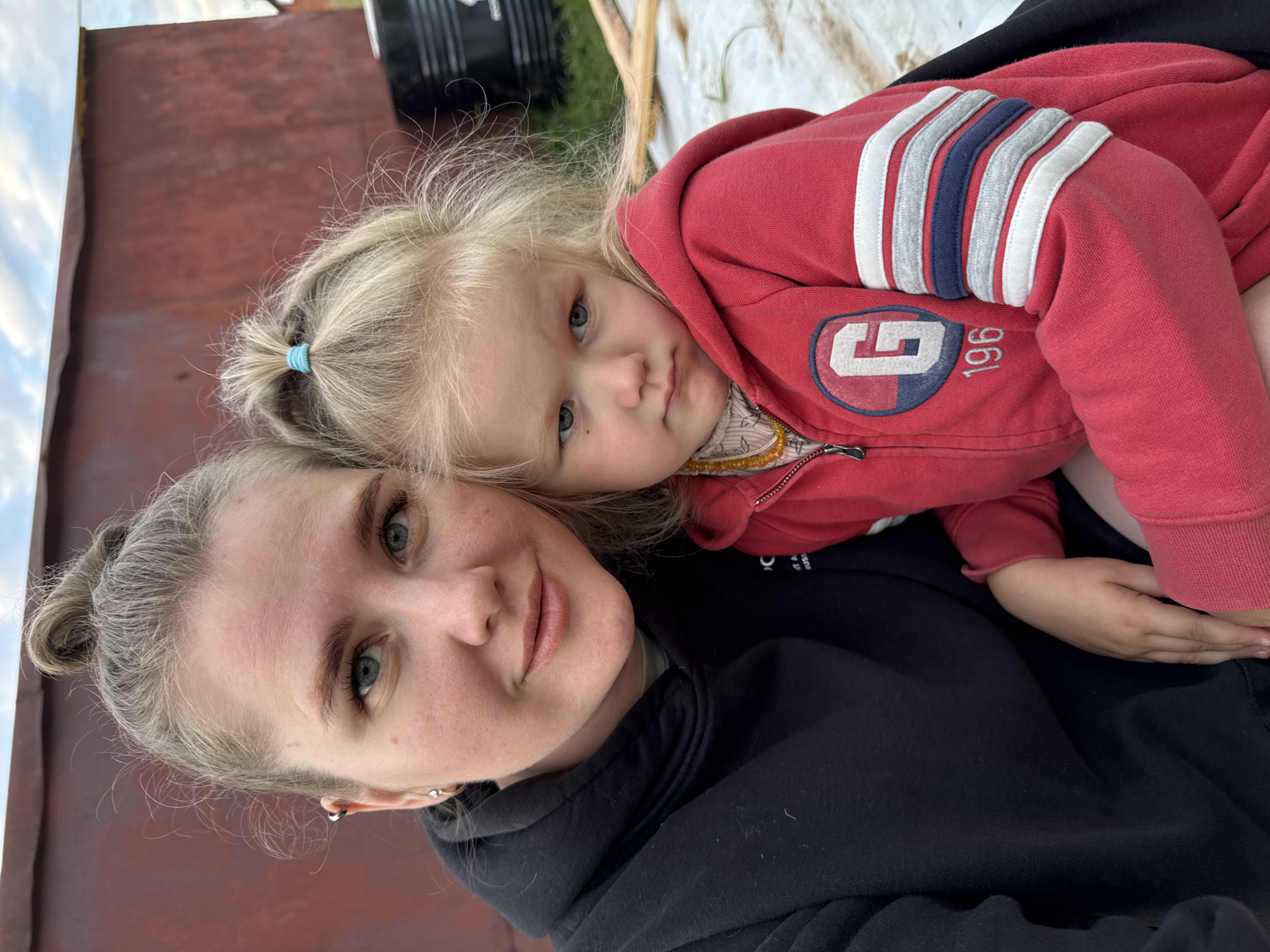 A woman and a young child are shown in a close-up, vertically oriented photograph. The woman, with her hair pulled back and wearing a black hoodie, is looking directly at the camera. The child, with blonde hair tied in a small ponytail, is resting on the woman's shoulder, looking towards the camera with a soft expression. The child is wearing a red hooded jacket with white and blue stripes on the sleeves and a circular emblem on the chest containing the letters "G" and the number "196". The background is blurred, showing a reddish-brown surface and some green elements, possibly foliage. The lighting suggests it is daytime, likely outdoors. The overall mood appears intimate and affectionate, with both subjects displaying calm and contented emotions. The visible text on the child's jacket is "G 196". The location is identified as a farm in Russia.