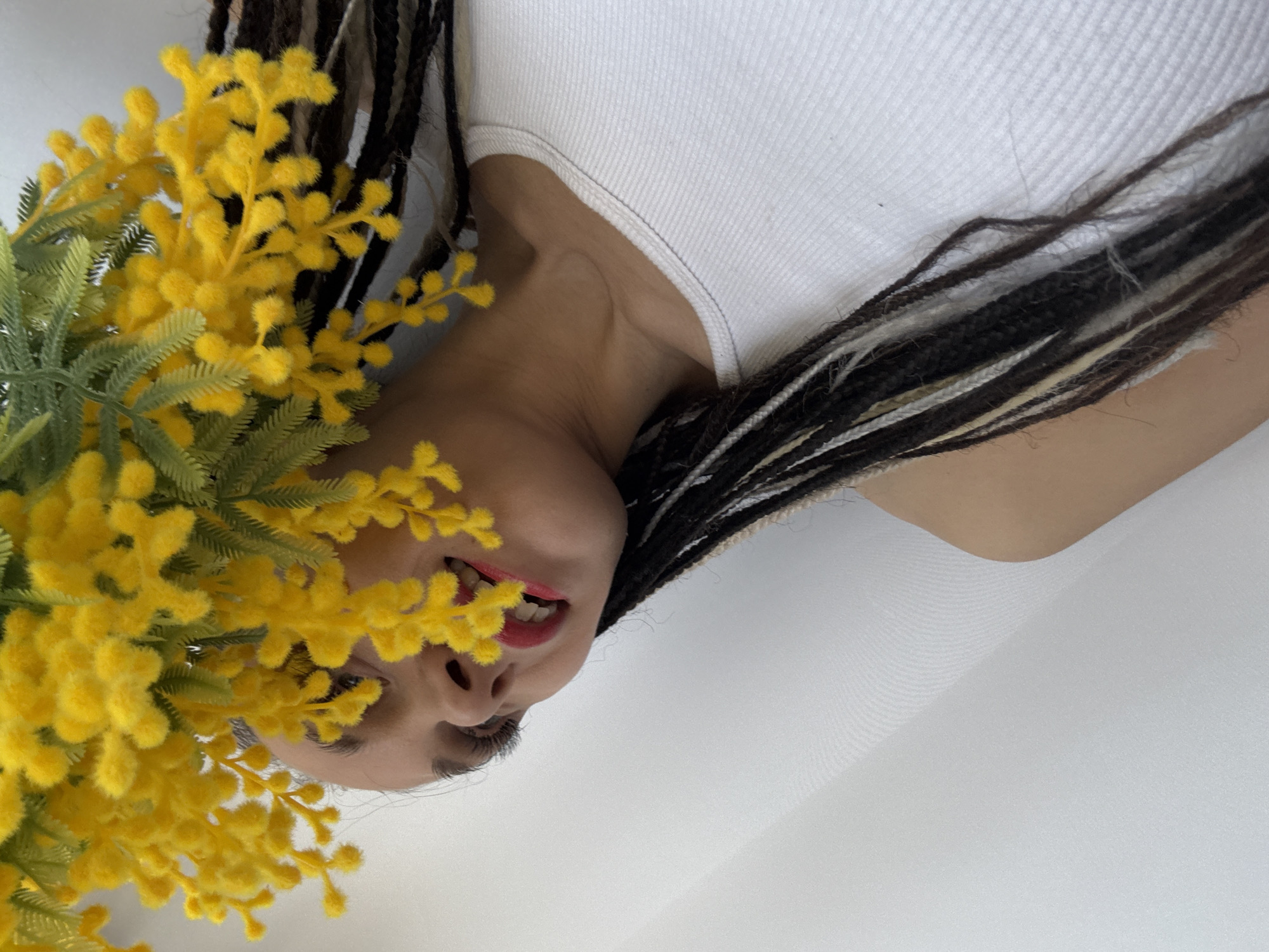 A woman wearing a white tank top lies with her head tilted downwards, framed by yellow flowers on the left and her dark braids on the right. The background appears to be a white surface. Her face is visible, and she has red lipstick on.