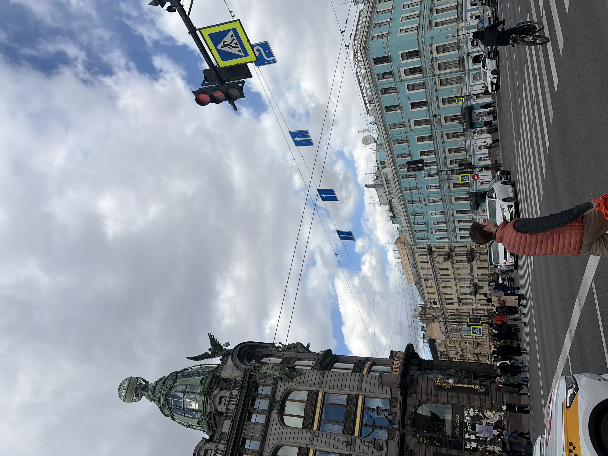 This image, rotated 90 degrees clockwise, presents a dynamic street scene in Saint Petersburg, Russia, under an overcast sky. The view looks up from street level towards the architectural details of two prominent buildings and the sky above.
On the left side of the street, the iconic Singer House (Dom Knigi) stands out with its intricate Art Nouveau architecture. Its distinctive green-patinated dome, topped with a globe and sculptures of eagles, is a focal point against the cloudy backdrop. The building's ornate facade features arched windows and gilded accents, with the Cyrillic word "КОМПАНИЯ" (KOMPANIYA) visible on an upper part and "cafe René" on a shop window at street level.
Opposite, on the right side of the street, a light blue multi-story building with numerous windows stretches along the block. The street separating these buildings features a clearly marked pedestrian zebra crossing. A crowd of pedestrians is gathered at the crossing, some waiting, others potentially in motion. In the immediate foreground, a person in a reddish-orange puffer vest is partially visible. Further down the street, a cyclist is seen riding.
Overhead, traffic infrastructure includes a red traffic light and multiple street signs. A prominent yellow square sign with a blue triangle indicates a pedestrian crossing, and several blue rectangular signs with white arrows direct traffic. Overhead wires crisscross the sky. The daytime scene is characterized by soft, diffuse light due to the heavy cloud cover.