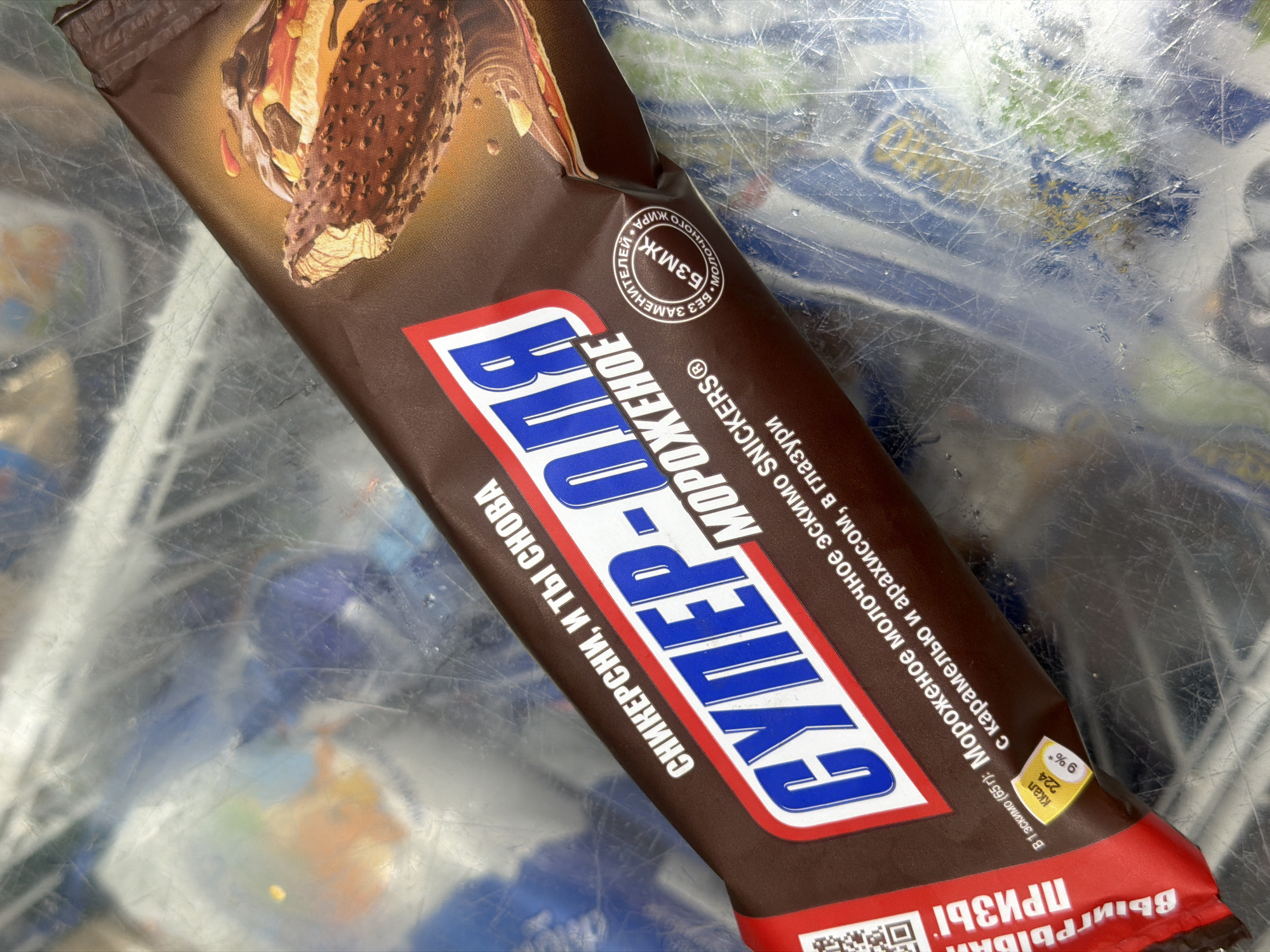 The image shows a package of Snickers ice cream, specifically "Super-Ola Ice Cream." The packaging is brown and features an image of the ice cream product. The text on the package is in Russian, indicating the brand, flavor description, and nutritional information. The package rests on a patterned surface, with what appear to be scratches on it. The picture was probably taken indoors, perhaps at a store or home.