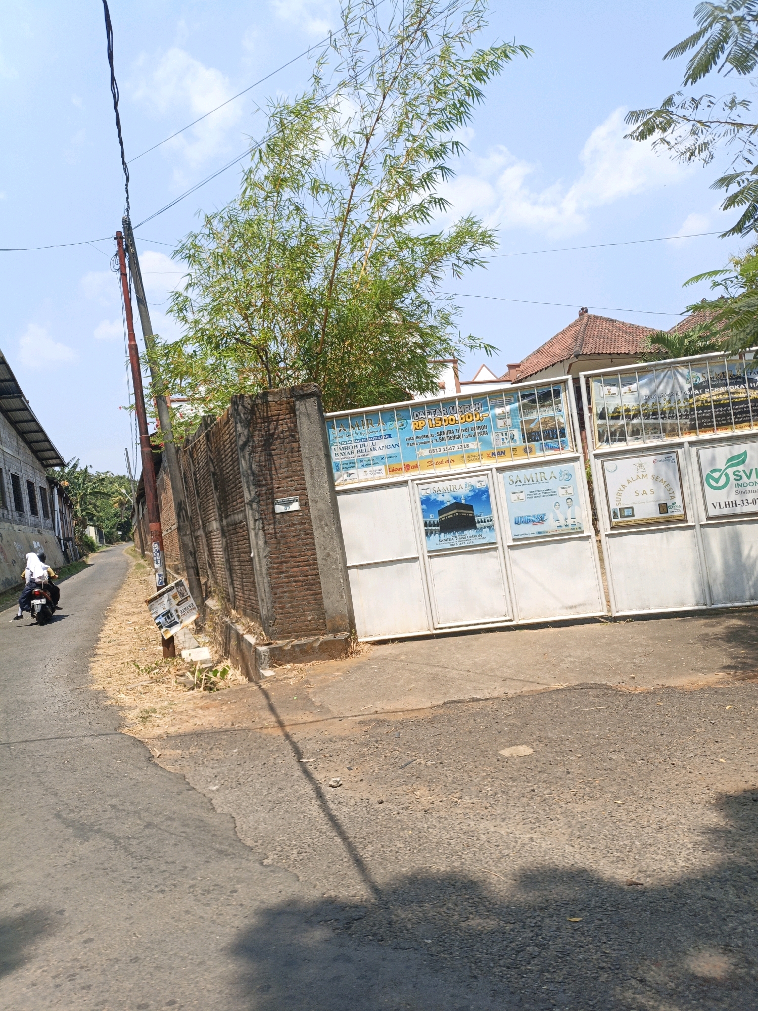 This is a photograph taken on a sunny day in Bandengan, Indonesia. The image depicts a street scene with a few key elements. In the foreground, a paved road is visible, with a slight tilt to the left. To the right of the road, there is a concrete wall with a large, white metal gate. Several posters are affixed to the gate, advertising what appear to be travel or religious services, including one with an image of the Kaaba. Above the gate, bamboo plants extend into the frame, their thin leaves reaching towards the bright blue sky with scattered white clouds.
To the left of the road, there is a row of buildings with corrugated metal roofs and windows. Further down the street, a person wearing a white hijab and yellow shirt is riding a motorcycle, heading away from the camera. A power pole with wires runs along the side of the road. The ground beside the road is covered with dry grass and some debris, and a signpost with a tilted sign displaying the word "ESTATE" is visible near the gate. The overall impression is of a local street with some commercial or service-oriented establishments.