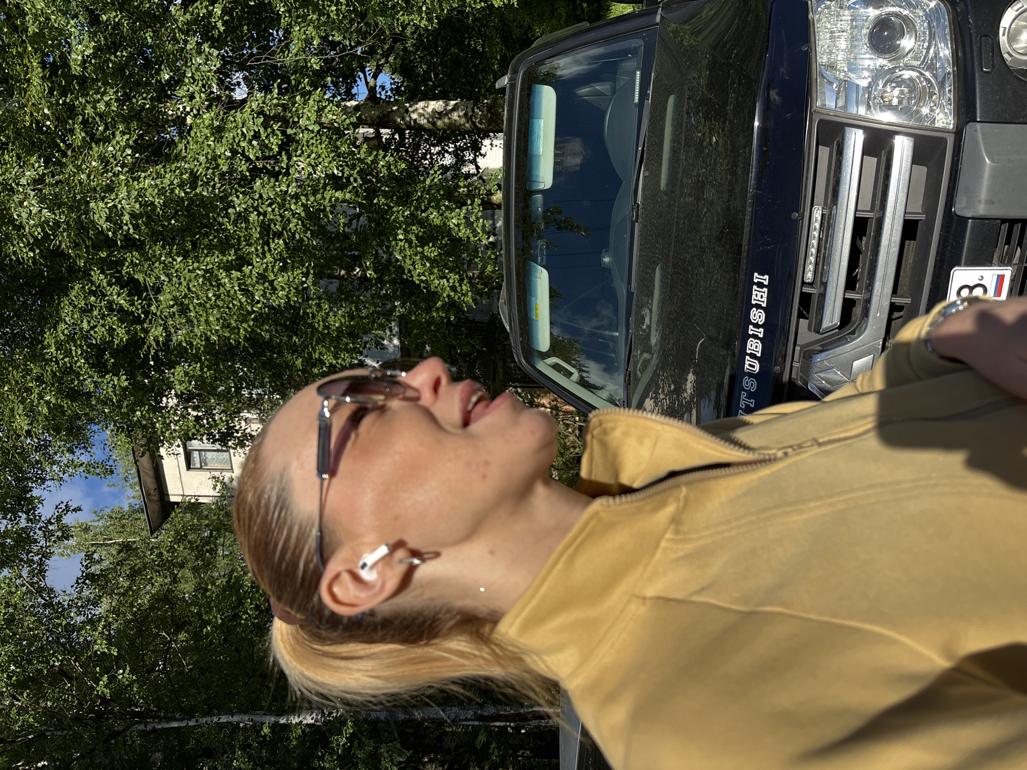 A close-up shot focuses on a woman in profile, facing left, as she stands in front of a black Mitsubishi Pajero. She has blonde hair pulled back in a ponytail, wears sunglasses, and has white earbuds in her ears. She is wearing a yellow jacket, and her left arm, wearing a watch, is visible in the foreground. The car's grille and headlights are clearly visible, and a license plate with Russian markings is on the bumper. Behind the car, lush green trees and a white building with windows can be seen under a bright sky. The scene appears to be outdoors, possibly in a residential area of a city in Russia, given the car and building details.