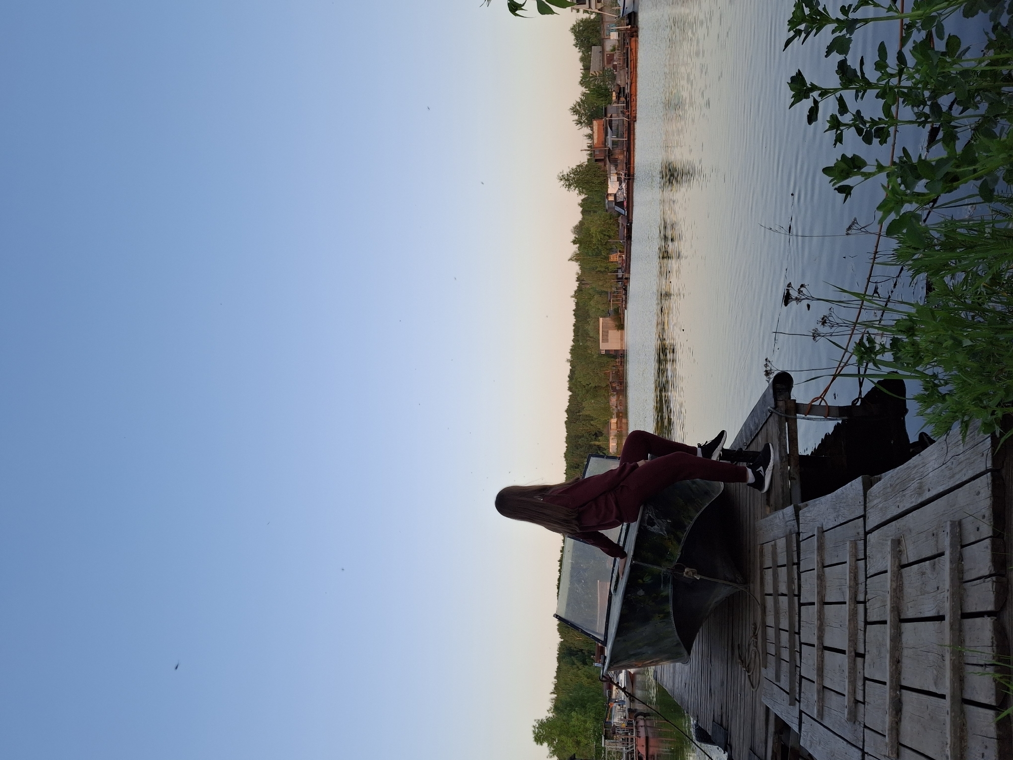 A photograph depicts a person with long hair, wearing a maroon sweat suit, seated on the edge of a dark-colored boat located on a wooden pier. The person is facing away from the camera, towards a body of water.
The pier is made of weathered wooden planks. The boat is partly submerged in the water.
Across the water is a shoreline with trees and what appears to be buildings, likely a residential area.
The sky is a gradient of light blue to pale orange, suggesting dawn or dusk.
The location is identified as Chaikovsky, Russia, based on information provided. Vegetation is visible on the right side of the pier.