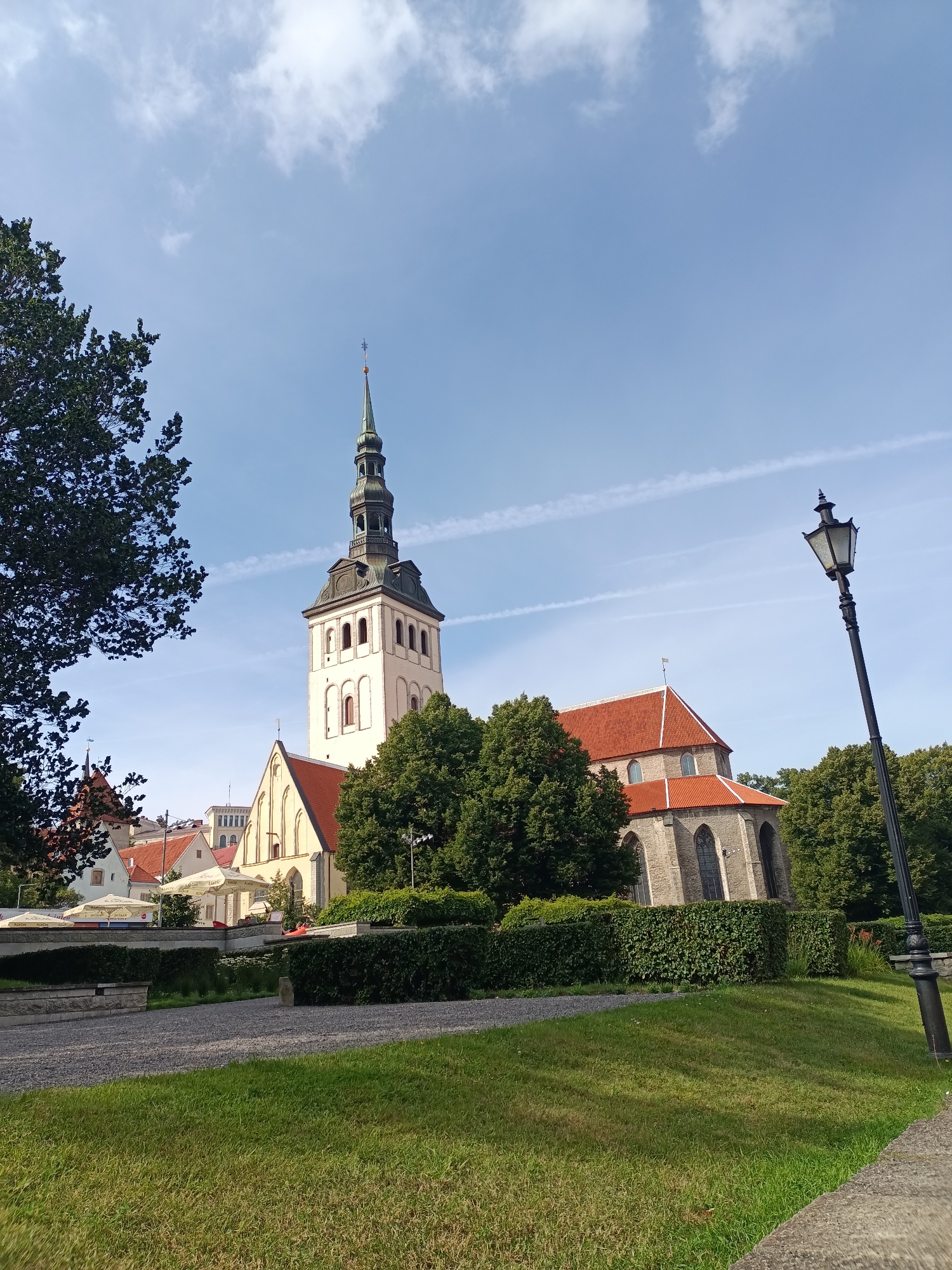 The image captures a scene in Tallinn, Estonia, featuring the iconic St. Olaf's Church (Oleviste kirik). The church's tall, slender white tower with a green spire dominates the background, set against a bright blue sky with wispy clouds. The church's nave, with its red tiled roof and stone walls, is visible to the right of the tower. Lush green trees and meticulously trimmed hedges surround the church, creating a park-like atmosphere. A cobblestone path runs through the foreground, and a vintage black lamppost stands tall on the right side of the frame. In the mid-ground, a row of buildings with red roofs and awnings suggest a commercial area or cafe. The overall impression is one of a well-preserved historical area on a sunny day.