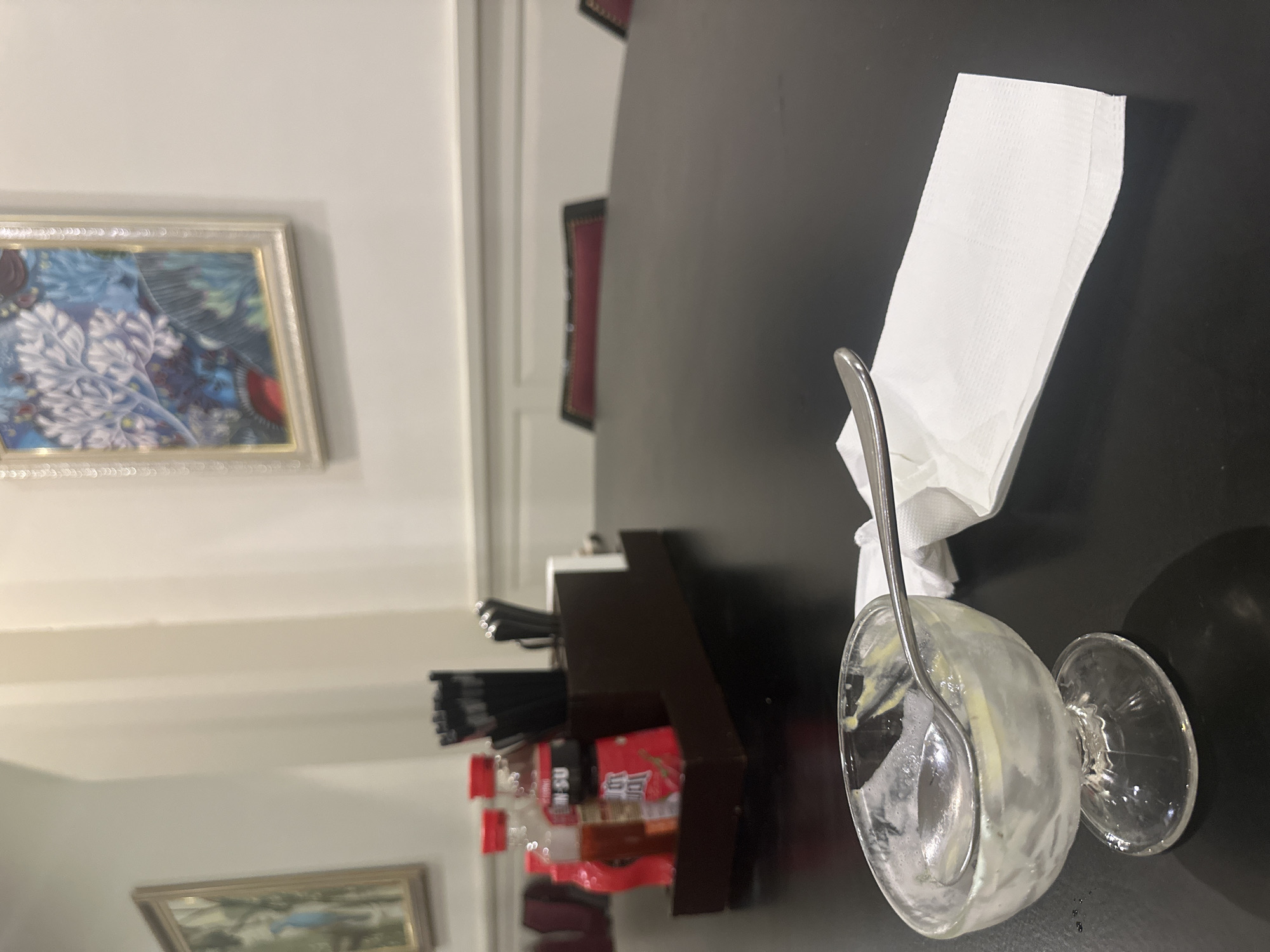 A photograph shows a table setting with a partially eaten dessert in a clear glass bowl with a spoon resting inside. A white napkin is crumpled next to the bowl. To the side, there is a holder containing utensils and bottles of condiments. In the background, there is an ornate wall with framed artwork. The location is in Unknown, Unknown.