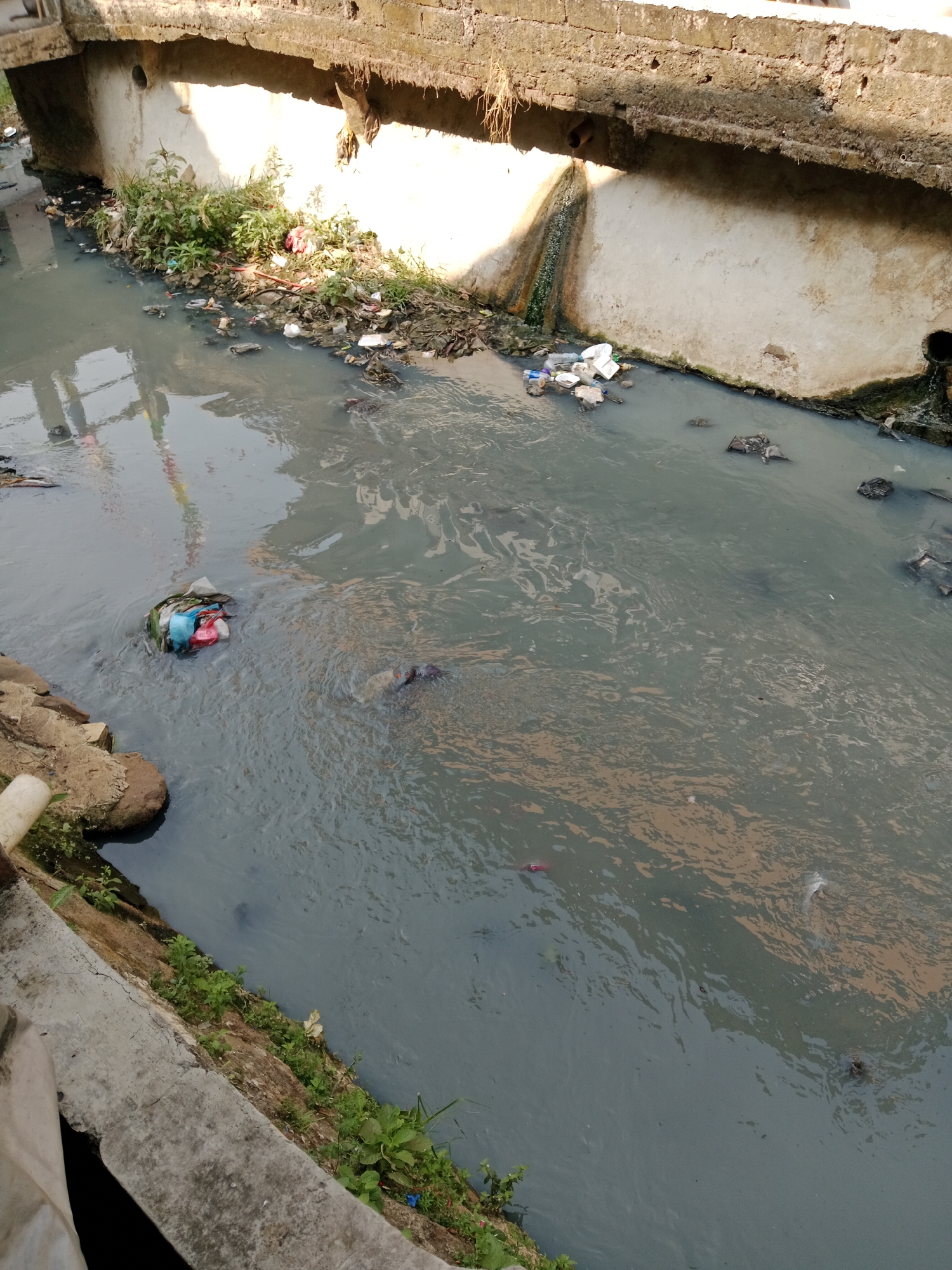 The image depicts a polluted canal in East Jakarta, Indonesia, characterized by murky, dark water filled with visible debris and waste. A concrete bridge or overpass spans the canal at the top of the frame, with its underside showing signs of decay and staining from the water. Along the canal banks, particularly on the left, there is an accumulation of garbage, including plastic bags, bottles, and other refuse, interspersed with green vegetation. The water itself contains floating trash, including a clump of plastic bags and other materials in the mid-left of the image. Further down the canal, more waste is visible, some submerged and others floating on the surface. The overall impression is one of severe environmental pollution and neglect.