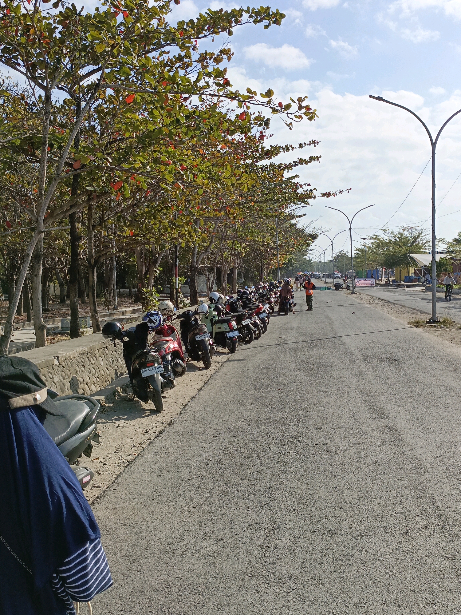 The image depicts a roadside scene in Bulukumba, Indonesia, likely near a coastal area given the presence of palm trees and the general ambiance. A row of parked motorcycles lines the left side of the asphalt road, angled slightly towards the viewer. The motorcycles are of various colors and models, some with helmets resting on them. To the left of the motorcycles, a low stone wall separates the road from a sandy area covered with dried leaves. Behind the stone wall, a dense line of trees with autumn-colored leaves forms a natural canopy over the area.
The road stretches into the distance, curving gently to the right. Several streetlights with curved poles are visible along the road, suggesting it's a well-lit area. In the middle distance, a person in a bright orange vest stands near the motorcycles, appearing to be directing traffic or overseeing the parking area. Further down the road, there are more parked vehicles, some buildings with awnings, and other pedestrians. The sky is blue with scattered white clouds, indicating a bright, sunny day. In the foreground on the far left, the back of a person wearing a dark blue garment and a black cap is partially visible, facing away from the camera. The overall scene suggests a public area, possibly a tourist spot or a marketplace, with a moderate amount of activity.
