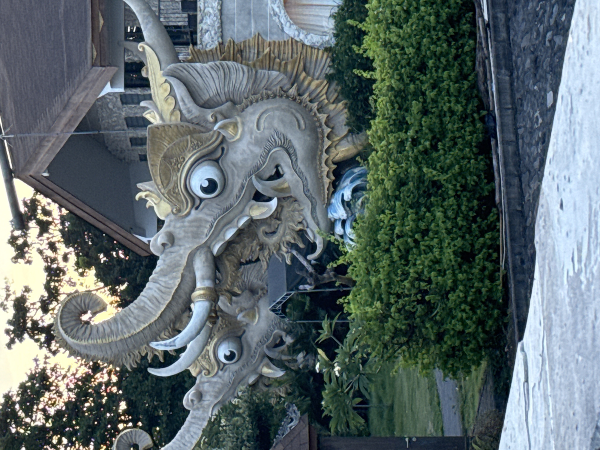 The image captures a monument featuring two intricately designed elephant-like creatures with prominent tusks and large, expressive eyes. The monument is located near a building and is partially surrounded by lush greenery, including bushes and trees. The scene is bathed in natural light. The location of the monument is in the city of Denpasar, Indonesia.