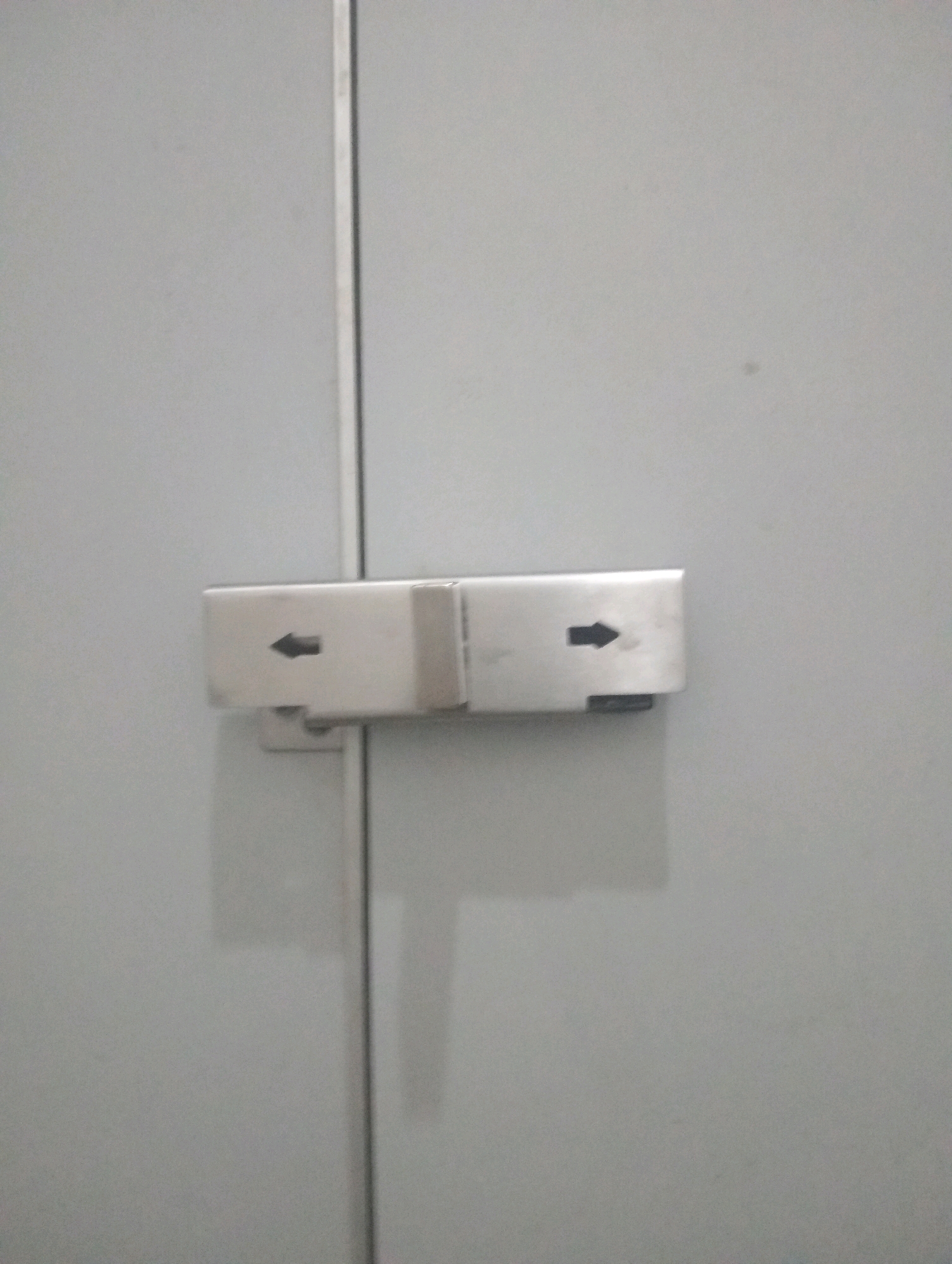 The image shows a close-up of a metal lock mechanism on a light gray surface, likely a door or cabinet. The lock is composed of two parts, each with a black arrow pointing in opposite directions. The left part of the lock has an arrow pointing left, and the right part has an arrow pointing right. The lock is situated in the city of Batu, Indonesia, suggesting it may be part of a public facility, such as a restroom or a locker. The environment appears clean and functional, with no visible people or other objects in the immediate vicinity.