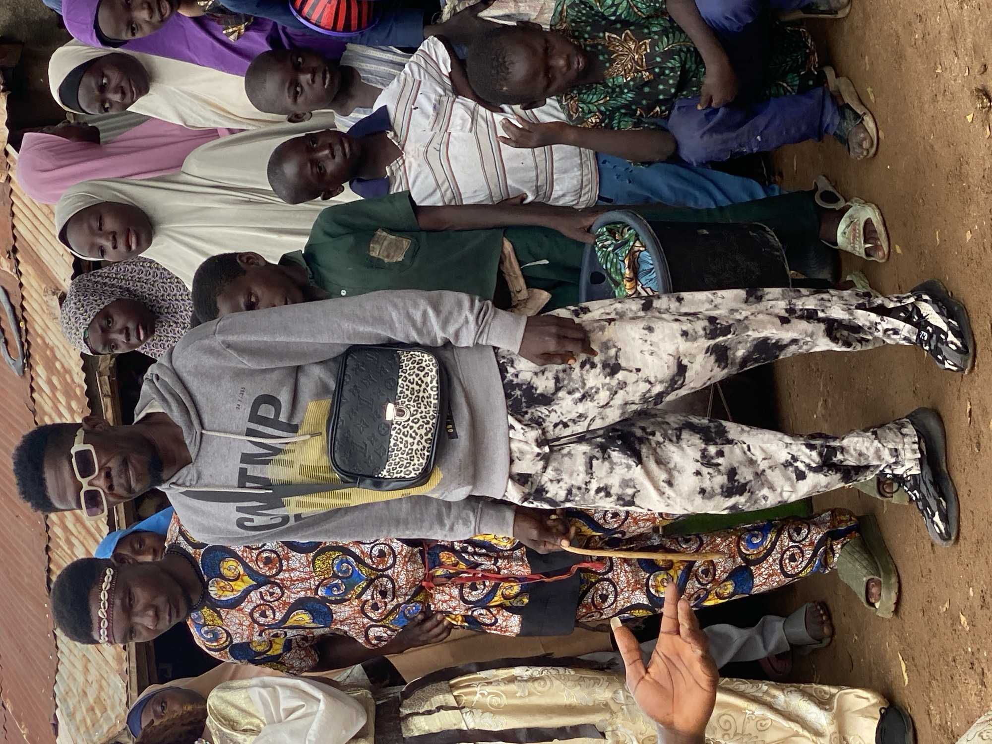 In the city of Rigachikun, Nigeria, a group of people are gathered outdoors, in front of a structure with a corrugated metal roof. The ground is composed of dry, brown soil.
The primary subject is a man standing prominently in the foreground, wearing a grey hooded sweatshirt and pants with a black and white marble pattern. He has a crossbody bag with a leopard-print front pocket. He wears sunglasses and a headband adorned with shells. He holds a thin wooden stick.
To his left, a man in a vibrantly patterned traditional outfit, featuring blue, yellow, and red motifs, stands with a similar headband. To the right of the primary subject, a group of children are present. One child, wearing a green patterned shirt and blue pants, is seated on the ground. Other children stand around him, some wearing t-shirts and others traditional attire. Several women, some wearing hijabs in various colors including purple, light pink, and cream, are visible in the background. The overall scene appears to be a social gathering or community event.