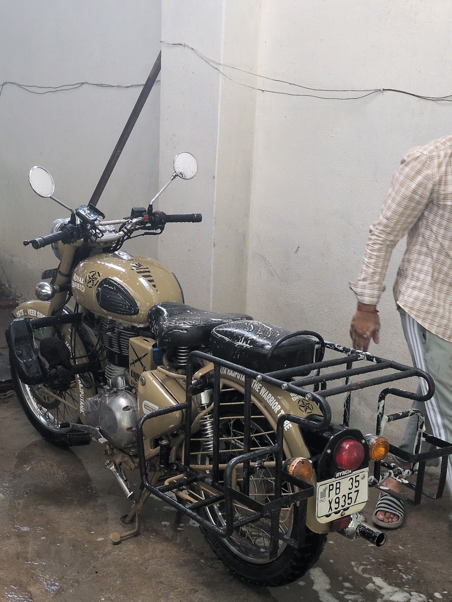 The image depicts a Royal Enfield motorcycle parked on a wet concrete floor, possibly indicating it is being washed. The motorcycle is light brown with black accents and features various accessories, including a luggage rack and custom detailing on the fuel tank and rear fender. The license plate reads "PB 35 X9357". A person, wearing a checkered shirt and light-colored pants with stripes, is partially visible on the right side of the frame, with their hand extended towards the rear of the motorcycle. The background consists of a textured, off-white wall with some wires running along it. The overall scene suggests an outdoor or semi-outdoor setting, possibly a service area or a residential courtyard, given the urban infrastructure implied by the wall and the motorcycle's presence. The specific location is the city of Pathankot, India.