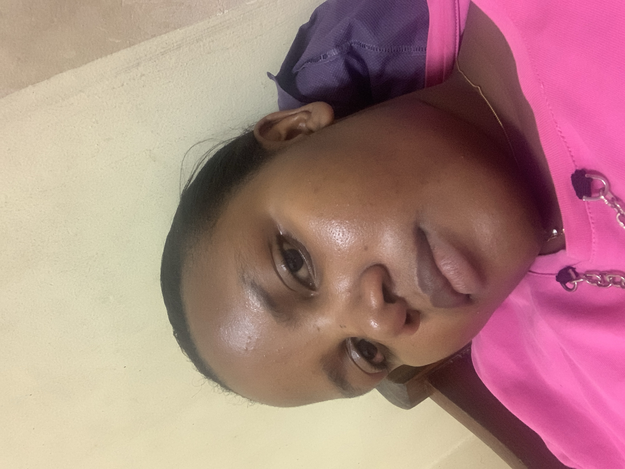 A close-up, rotated portrait depicts a person with dark hair and skin. The person is wearing a bright pink garment with silver chain details and a gold necklace. A purple bag is positioned behind their head, resting on a light yellow wall. The setting cannot be determined with enough confidence to assert that it is located in Dar es-Salaam, Tanzania.