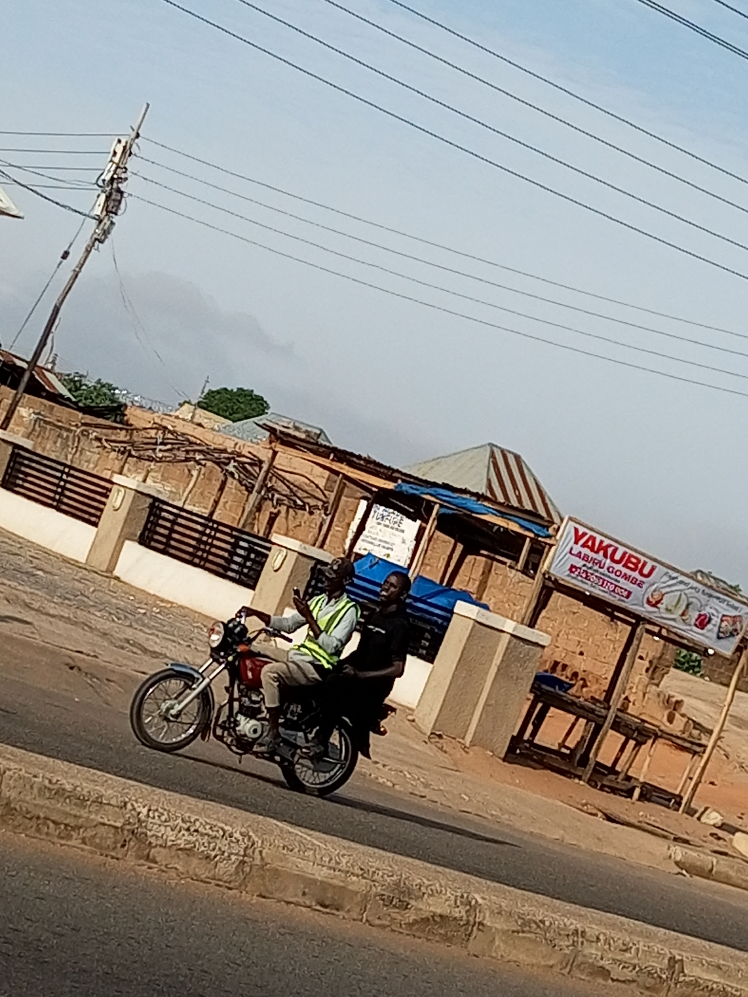 The image depicts a street scene in Garko, Nigeria. Two individuals are riding on a motorcycle. The person in front is wearing a reflective vest and appears to be holding a mobile phone. The person behind them is wearing a dark shirt. They are traveling on a paved road with a textured curb. In the background, there is a building with a sign that reads "YAKUBU LABIPU GOMBE." Various structures, including stalls and overhead wires, are visible, suggesting a market or roadside commercial area. The sky is overcast.