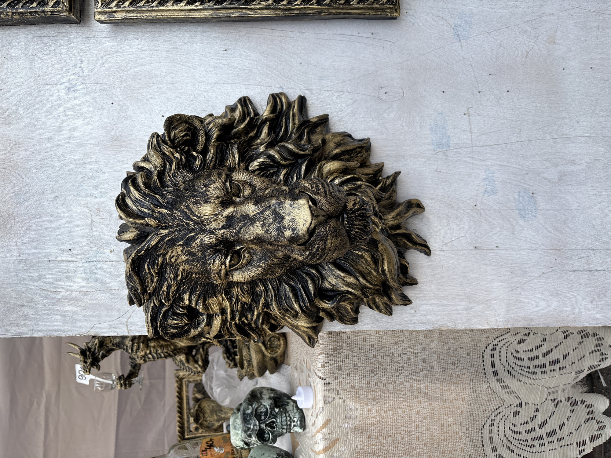 The image captures a close-up of a decorative lion's head sculpture mounted on a light-colored wooden wall. The sculpture is detailed, with a textured mane and face painted in a gold and black finish. To the left of the lion's head, a portion of a display table is visible, covered with a beige lace tablecloth. On the table, several other decorative items are present, including a skull-shaped object, a small white LED candle, and a framed picture. Further in the background, a statue of a dragon holding a glass with a price tag indicating "90" can be seen. The overall setting appears to be an outdoor market or craft stall. There is no explicit indication of the location being Vitsebsk, Belarus, based solely on the visual information provided in the image.