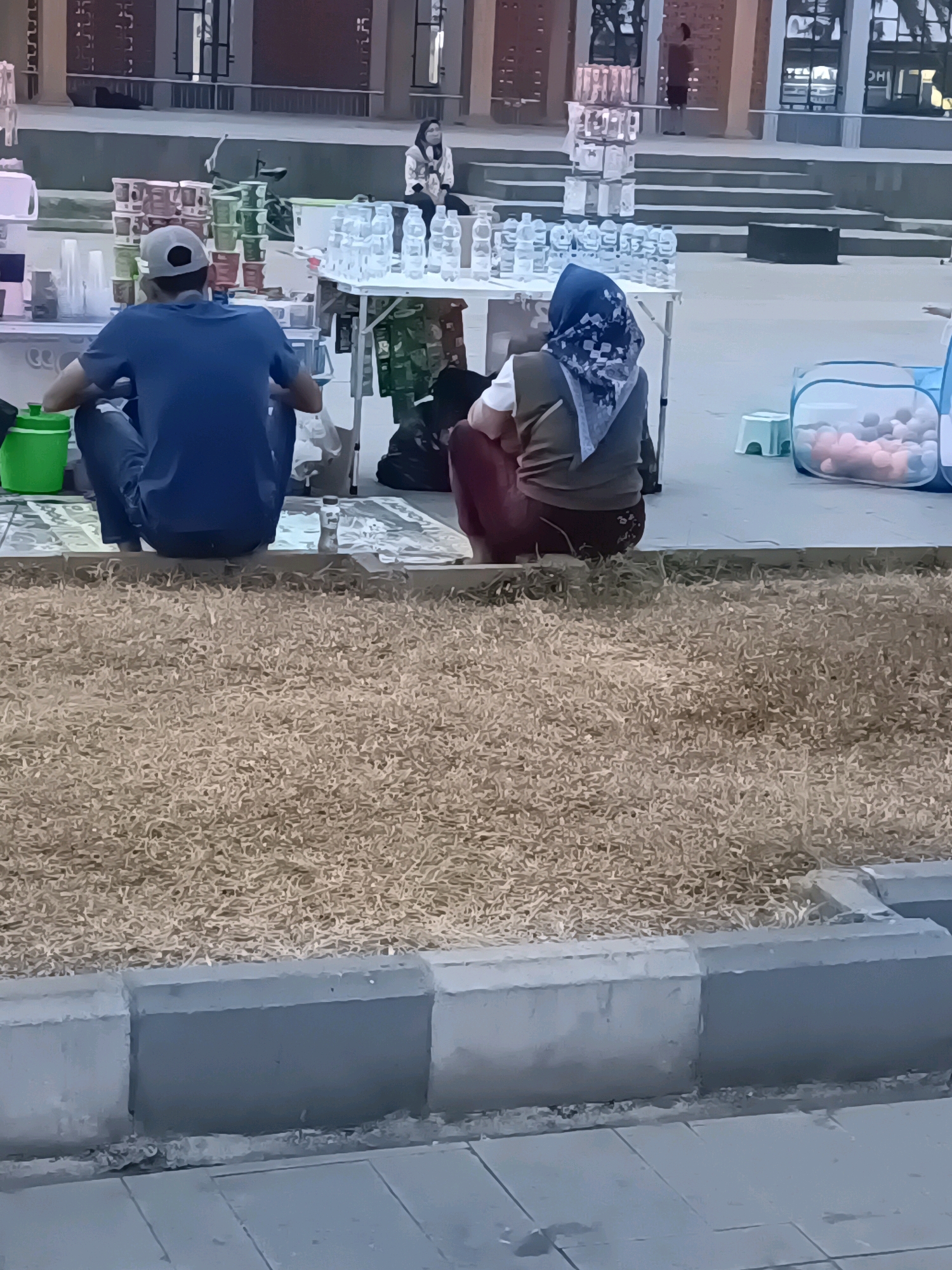 Two individuals are seated on the ground in front of a merchandise stall, facing away from the camera. The stall appears to be selling bottled water and other drinks. The person on the left is wearing a blue shirt and a grey baseball cap, while the person on the right is wearing a brown jacket and a patterned headscarf. In the background, a few more people are visible, along with steps and a building. The setting is outdoors, with dry grass and concrete structures in the foreground. The lighting suggests it might be dusk or early morning. This scene is consistent with street vending or informal market activities, which are common in public spaces in cities like Lubuklinggau, Indonesia.