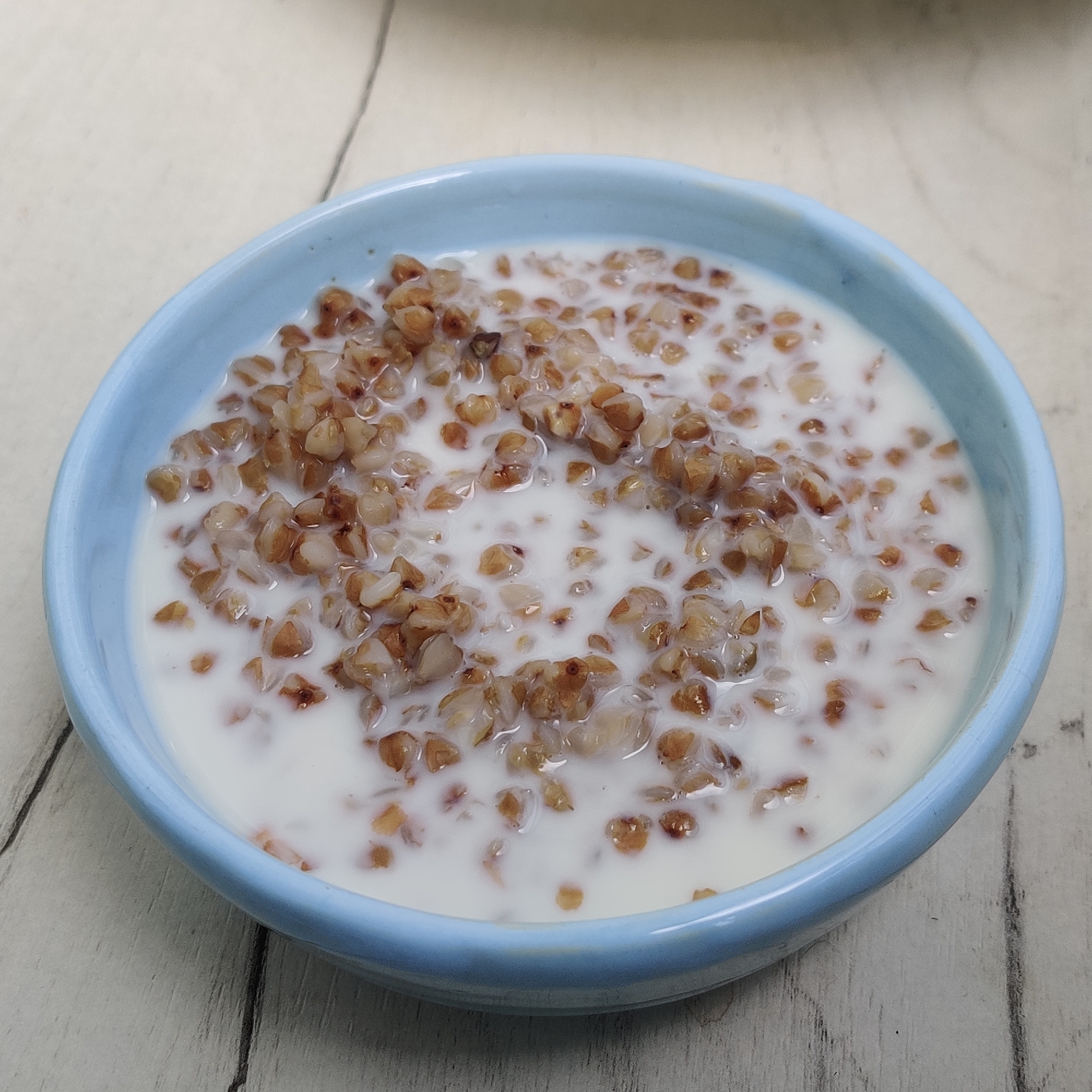 A close-up, overhead shot shows a light blue bowl filled with buckwheat porridge and milk. The porridge is a light brown color and the milk is white, creating a contrasting mix of colors within the bowl. The bowl is placed on a light-colored wooden surface, likely a table or countertop, with visible wood grain and subtle variations in color that suggest wear or a natural finish. The lighting appears to be soft and diffused, highlighting the textures of the food and the wood. There are no people or other objects visible in the frame. The scene is static and focuses solely on the bowl of food. The location in Yekaterinburg, Russia, is not discernible from the visual information provided in the image.