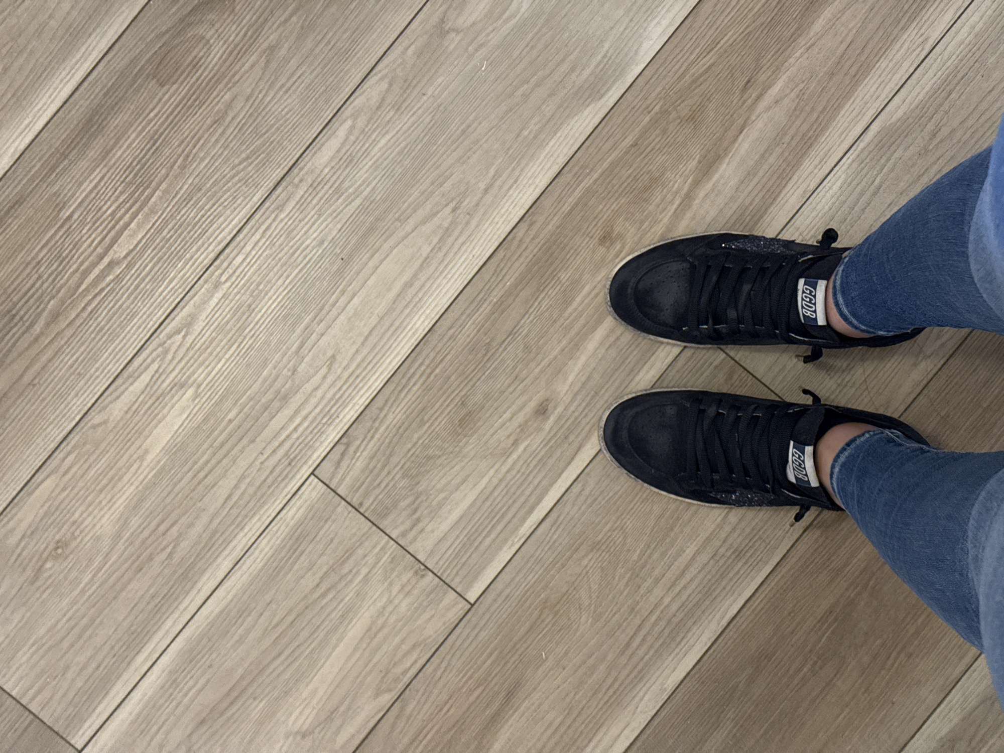 A first-person perspective image shows a pair of legs wearing blue jeans and black sneakers with the brand name "GGDB" visible on the sides, standing on a light brown wood-patterned floor. The floor planks are arranged at an angle. The location cannot be determined to be in San Antonio, United States of America, from the given information.