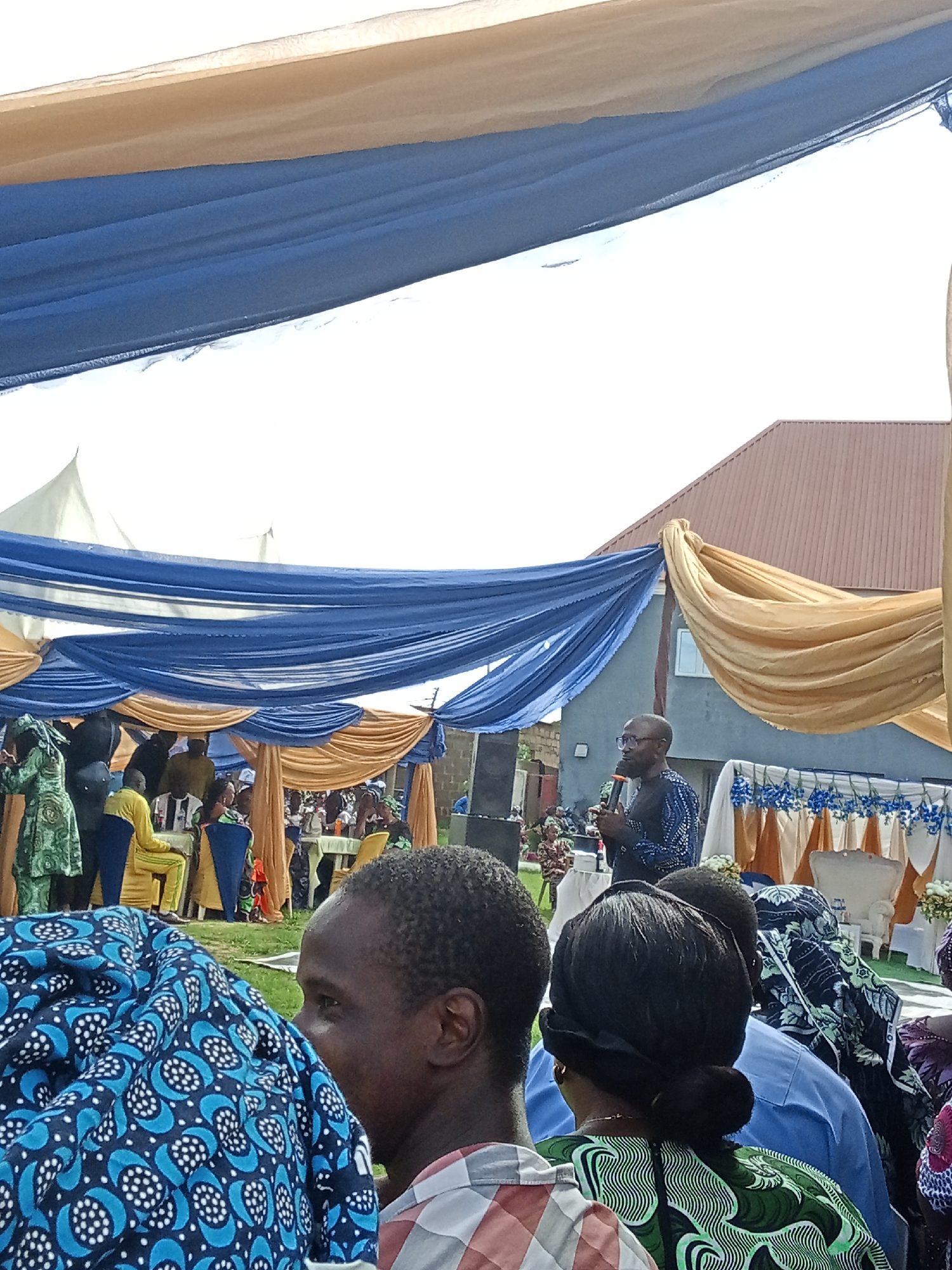 In Sabon Tasha, Nigeria, an outdoor event is taking place under a decorative canopy of draped blue and gold fabric. In the mid-ground, a man wearing glasses and a dark patterned shirt stands at a podium, holding a microphone and addressing an audience. The audience members are seated at tables in the background, with some people standing. In the foreground, the backs of several people are visible, dressed in various patterned fabrics. The overall scene suggests a social gathering or ceremony.