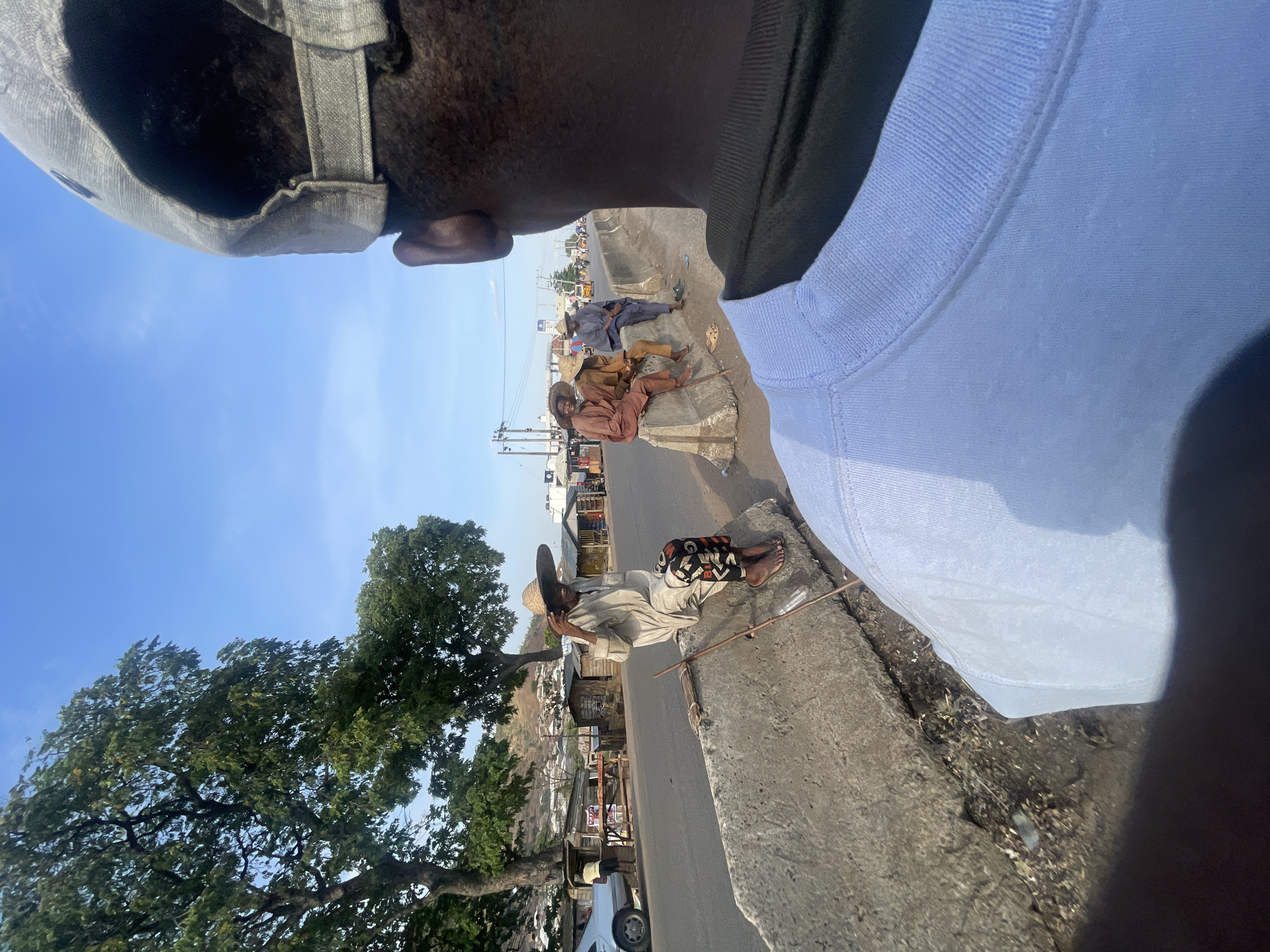 A street-level, partially obstructed view in Lokoja, Nigeria. In the foreground, three individuals are resting on a low concrete barrier that divides the pedestrian area from the road. The person closest to the camera is wearing a hat, light-colored clothing, and dark patterned leggings. They are leaning back, propped up by the barrier, with a wooden staff resting next to them. Further down the barrier, two more people are relaxing, dressed in traditional Nigerian attire. A large tree is growing beside the road, obscuring the shops. A car is partially visible near the tree. The sky is clear and blue. The image is taken from the point of view of another person whose head and shoulder are visible in the frame, suggesting the photo was taken with a cell phone.