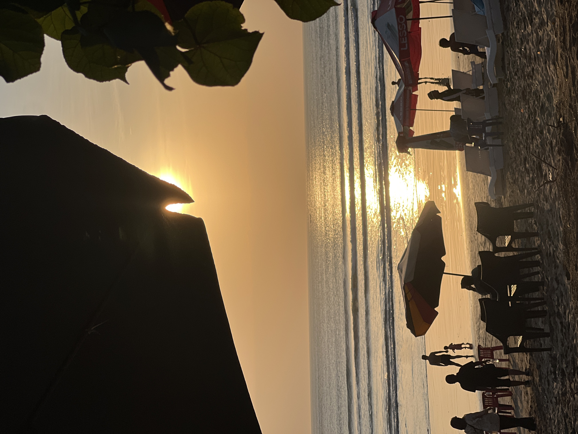 The image depicts a beach scene at sunset in Kuta, Indonesia. The sun is low in the sky, casting a warm, orange glow over the ocean and the beach. Gentle waves are rolling in, and their surface reflects the sunlight. In the foreground, the sandy beach is dotted with various objects and people. Several beach chairs and umbrellas are set up, with some occupied by silhouetted figures. Two red and white umbrellas with the word "LESSO" are visible, suggesting a commercial area. In the middle ground, more beach chairs and umbrellas are present. Several people are walking on the beach, including a man and a child in silhouette. The ocean extends to the horizon, where it meets the luminous sky. The scene is framed by dark silhouettes of foliage on the right and a large, dark structure or awning on the left, partially obscuring the top of the frame and the sky. The overall atmosphere is serene and tranquil, characteristic of a tropical sunset.