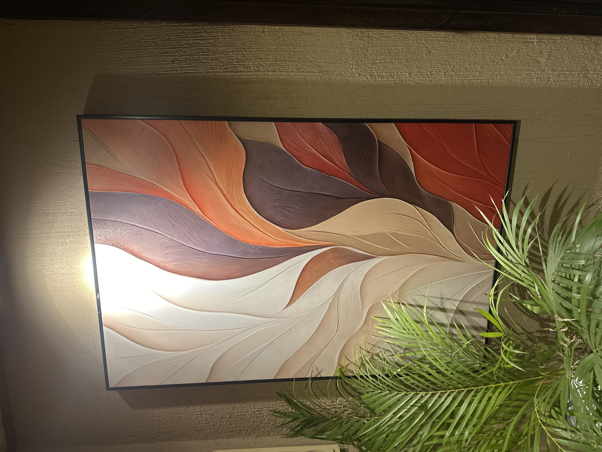 The image displays a framed abstract artwork on a textured wall. The artwork features overlapping leaf-like shapes in warm tones of white, beige, brown, and red. A potted green plant with feathery leaves is positioned in front of the lower left corner of the artwork, partially obscuring it. The wall itself has a rough, textured finish. Lighting from above casts a warm glow on the upper portion of the artwork and the wall. The scene appears to be an indoor setting, likely a home or commercial space. The location in Nam Hoa Lư Ward, Vietnam, is not visually indicated within the image itself.