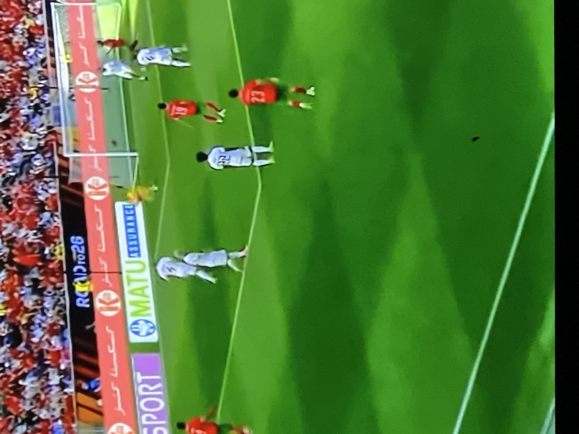 The image depicts a high-angle view of a soccer match occurring on a well-maintained green grass pitch, marked with white lines, within a stadium. The location is identifiable as a stadium in Casablanca, Morocco, based on the electronic advertising board displaying "MATU ASSURANCE," a Moroccan insurance company, and the text "ROAD to 26," referencing the 2026 FIFA World Cup which Morocco will co-host. Additional Arabic script is visible on other advertising banners and boards, including a prominent red banner with a 'K' logo behind the goal.
Approximately ten soccer players are visible on the field. One team wears white jerseys and shorts; a player with number '37' is centrally positioned, and another with number '3' is seen near the penalty area. The opposing team wears red jerseys and green shorts, with players numbered '19' and '23' visible in the mid-ground. A player in a yellow goalkeeper's kit is positioned near the white goal frame and netting on the right side of the frame. The players are in various active postures, indicating ongoing gameplay.
In the background, the stadium stands are filled with numerous spectators, predominantly wearing red and white attire. The scene is brightly lit, consistent with daylight or powerful stadium lighting. The image captures a specific moment during a live soccer event.