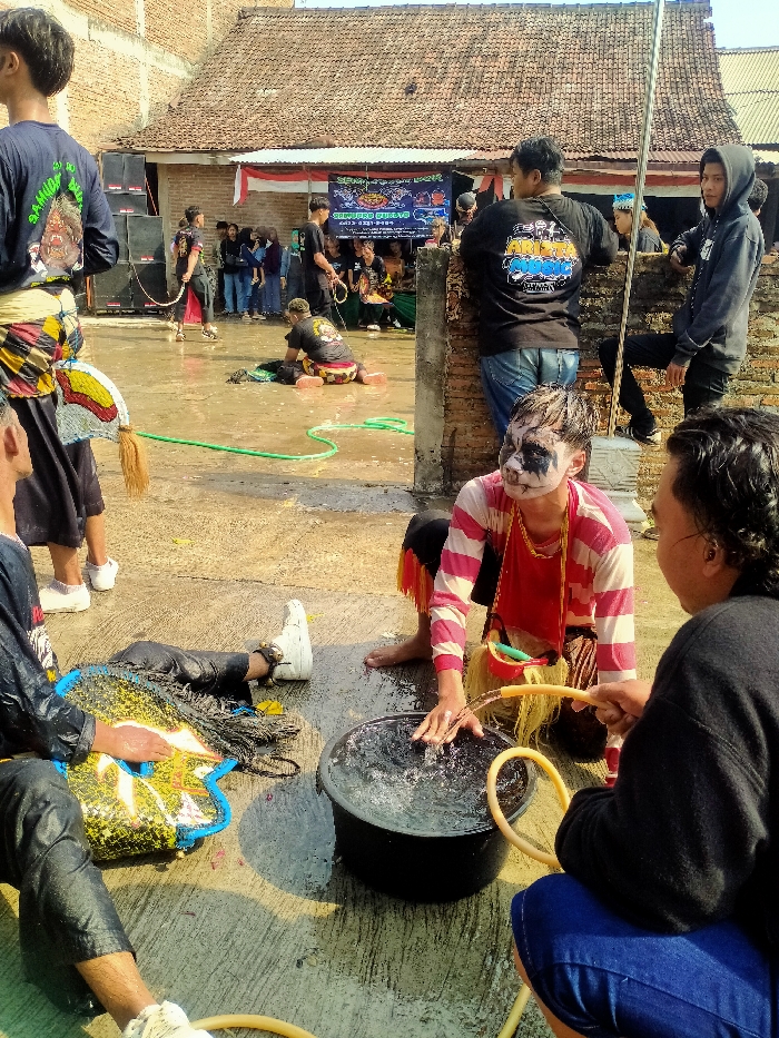 An outdoor scene in Sepat, Indonesia, features multiple individuals engaged in activities on a wet concrete surface under bright sunlight. In the foreground, a male individual with a white and black painted face, wearing a pink and white striped long-sleeve shirt and traditional attire with yellow fringe, kneels barefoot. He is immersing his right hand into a black bucket filled with water. Adjacent to him, a second male, seen from the back, squats while holding a yellow garden hose, directing water into the bucket. To the left foreground, a third individual lies supine on the wet ground, wearing dark pants and white athletic shoes, with a large, yellow and blue patterned object, consistent with a costume component, resting on their torso.
In the mid-ground, several other individuals are present. One male stands facing away from the viewer, wearing a dark long-sleeve shirt, a patterned waist sash, and white shoes. Further back and to the left, another male kneels on the wet ground near a dark object. Various other participants and observers are visible in the background, some standing, others leaning against a low brick wall on the right. The background features traditional Indonesian houses with terracotta tiled roofs and exposed brick walls. Large black sound speakers are positioned to the left. A banner with Indonesian text and red and white decorative flags are strung across the background, indicating an organized event.