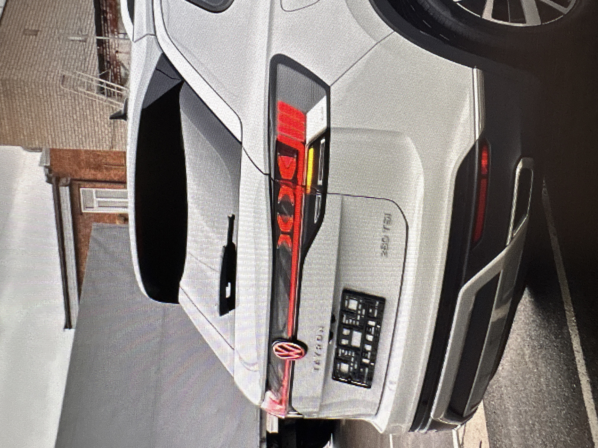 The image shows the rear passenger side of a white Volkswagen Tayron SUV parked on a wet asphalt surface, likely a street or parking lot, in Moscow, Russia. The vehicle's brake lights are illuminated, casting a red glow. A license plate holder is affixed to the rear bumper, but no license plate is visible. To the left of the SUV, a large, light-colored trailer or truck is partially visible. In the background, there are brick buildings and a cloudy sky. The overall lighting suggests it is daytime, possibly overcast.