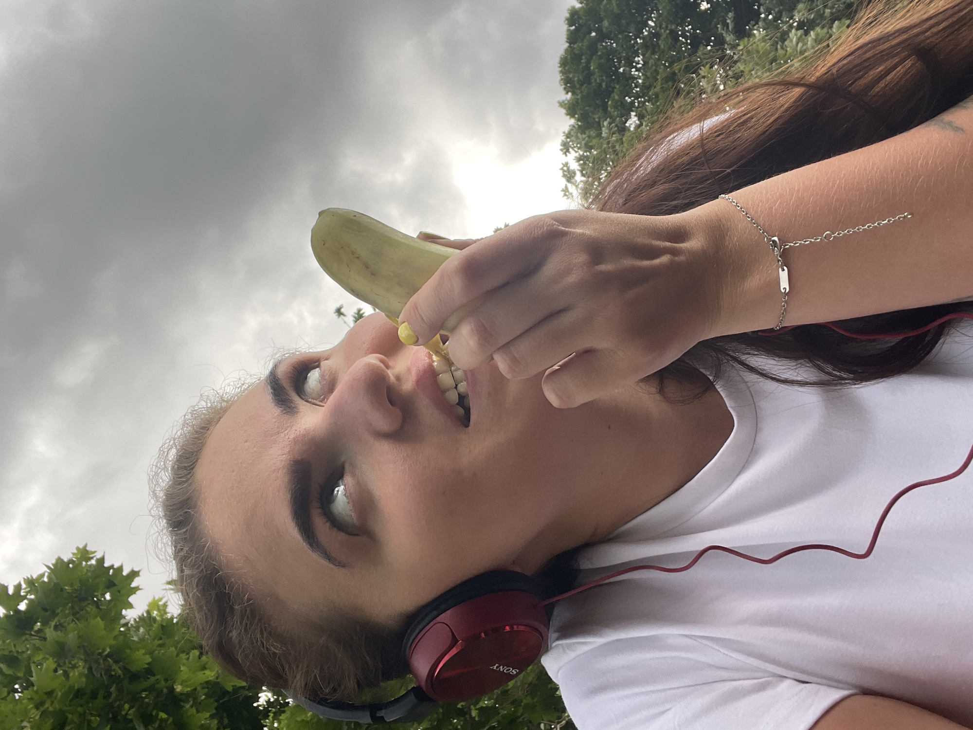 In a close-up shot, a young woman with brown hair, wearing red Sony headphones and a white t-shirt, looks upwards and to the right, holding a peeled banana to her mouth. Her eyes are wide and her mouth is open, with her teeth visible. She has bright yellow nail polish on her fingernails. A delicate silver bracelet with a chain dangle is visible on her left wrist. The background consists of green foliage and a cloudy sky. The scene appears to be outdoors in a natural setting, possibly a park or a wooded area in Ostrov, Russia, given the presence of trees and an overcast sky.