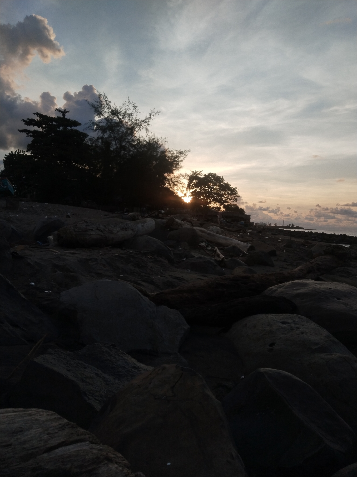 The image shows a sunset scene at a rocky beach in Lhokseumawe, Indonesia. Large, dark rocks dominate the foreground, leading up to a line of trees silhouetted against the bright setting sun. The sun is partially obscured by the trees, creating a burst of light. The sky is filled with clouds, some darker and more prominent to the left, while others are lighter and more diffused towards the right. In the distance, a strip of land and some structures are faintly visible. The overall tone of the image is dark due to the time of day and the focus on the shadowed rocks and trees.