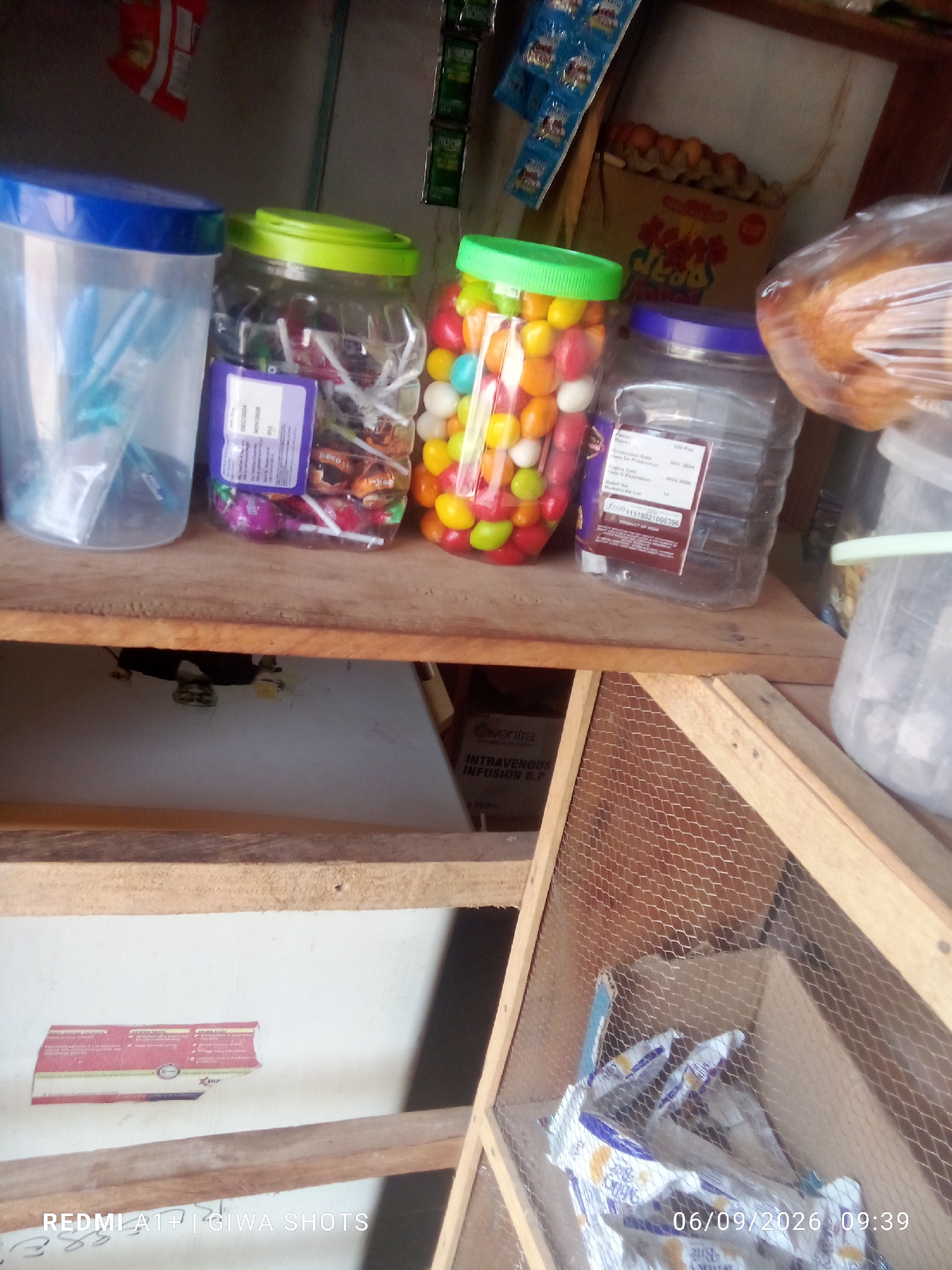 A cluttered, indoor market stall display in Jalingo, Nigeria features several plastic containers filled with candies and lollipops. On a wooden shelf, a clear plastic container with a blue lid holds blue plastic objects. Next to it, a clear plastic container with a lime green lid contains assorted lollipops. To its right, another clear plastic container with a red lid holds a mix of colorful round candies. Further to the right, a clear plastic container with a purple lid is partially visible. Behind these, a rack of small, foil-wrapped food items hangs from the wall. A loaf of bread wrapped in plastic is also visible to the right. Below the shelf, a white surface has a torn label with red and blue text. A section of wire mesh forms part of the display at the bottom right, obscuring some packaged goods. The overall lighting suggests an indoor setting with ambient light.