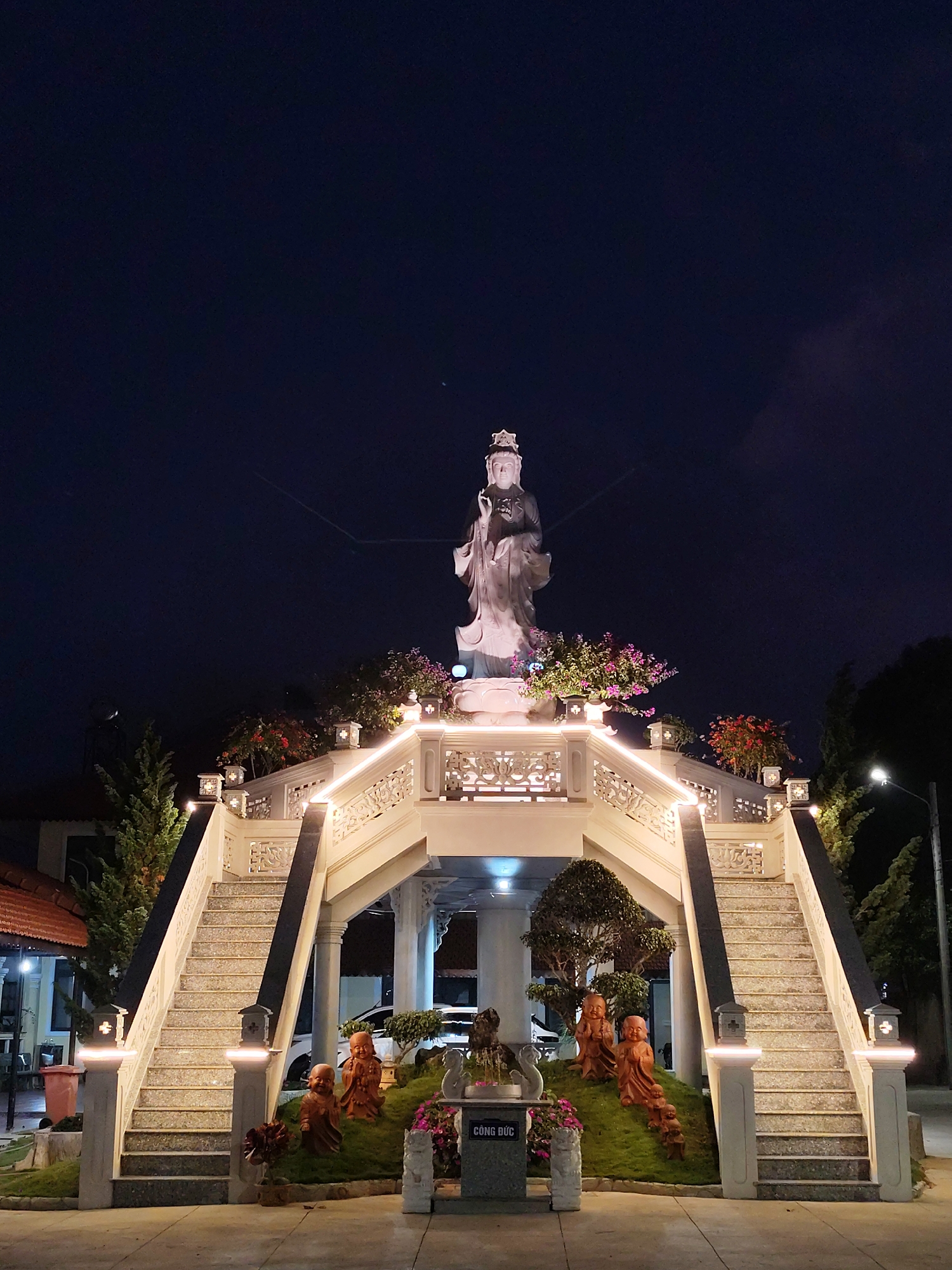 At night, the illuminated steps of a white stone bridge and archway lead to a white stone statue of a woman holding a plant at Linh Phuoc Pagoda in Lâm Viên - Đà Lạt, Vietnam. The archway has decorative lights and flower pots, and stairs go up either side. At the bottom are statues of small children in robes. “Công Đức” is printed on a gray slab at the base. A streetlight and trees are on the right, while another building is on the left. The sky is dark.