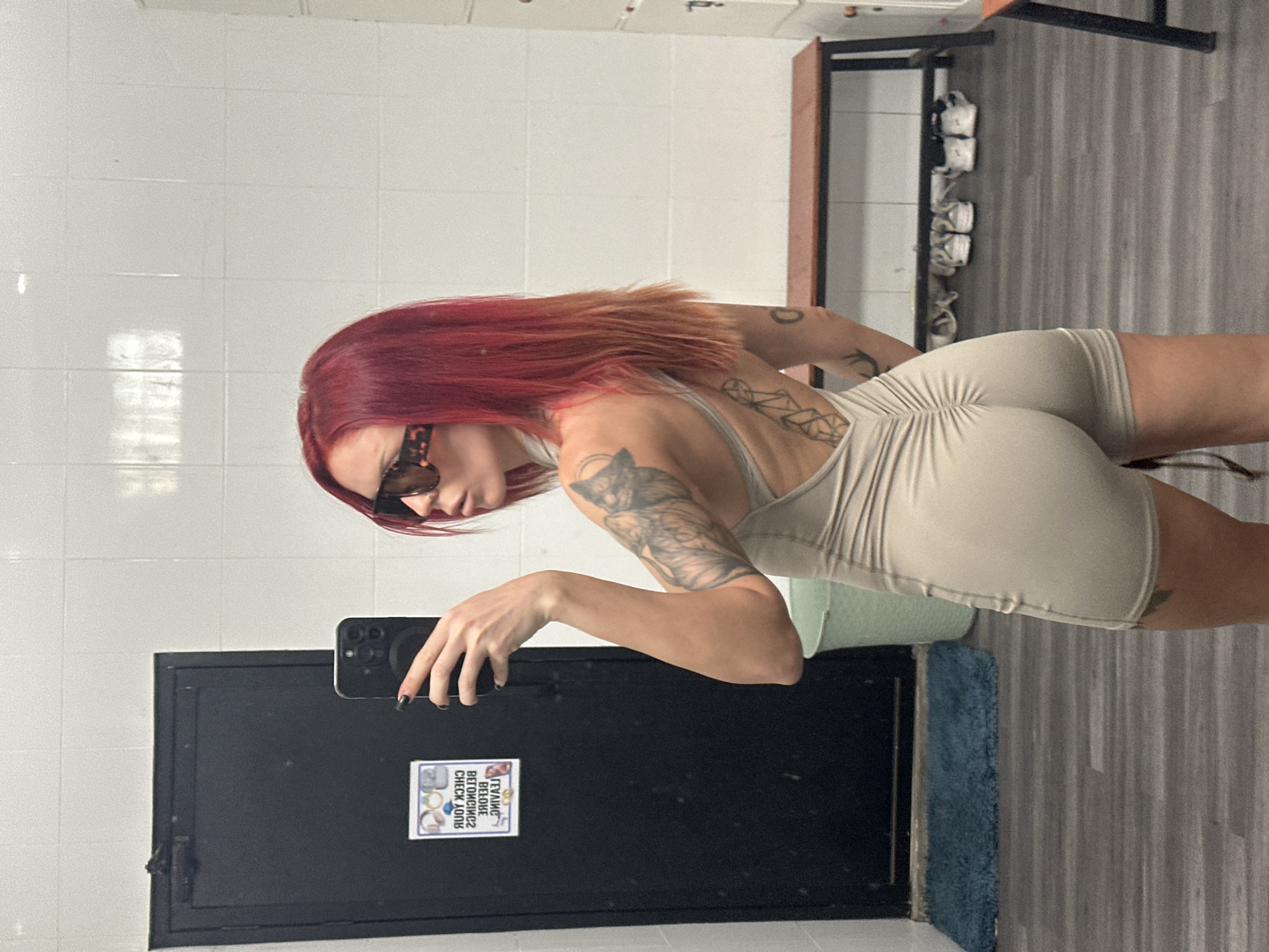 A woman with red hair, wearing a light-colored, form-fitting one-piece athletic outfit and sunglasses, is taking a selfie in a locker room. She is positioned with her back to the camera, looking over her shoulder at the mirror. Her left arm is raised, holding a smartphone. Tattoos are visible on her left arm and back. The locker room has white tiled walls, a dark door, a light green trash can, a wooden bench with lockers behind it, and a dark blue mat on the floor. The flooring appears to be wood laminate. There are several pairs of shoes lined up underneath the bench. The scene is indoors and appears to be a gym or similar facility.