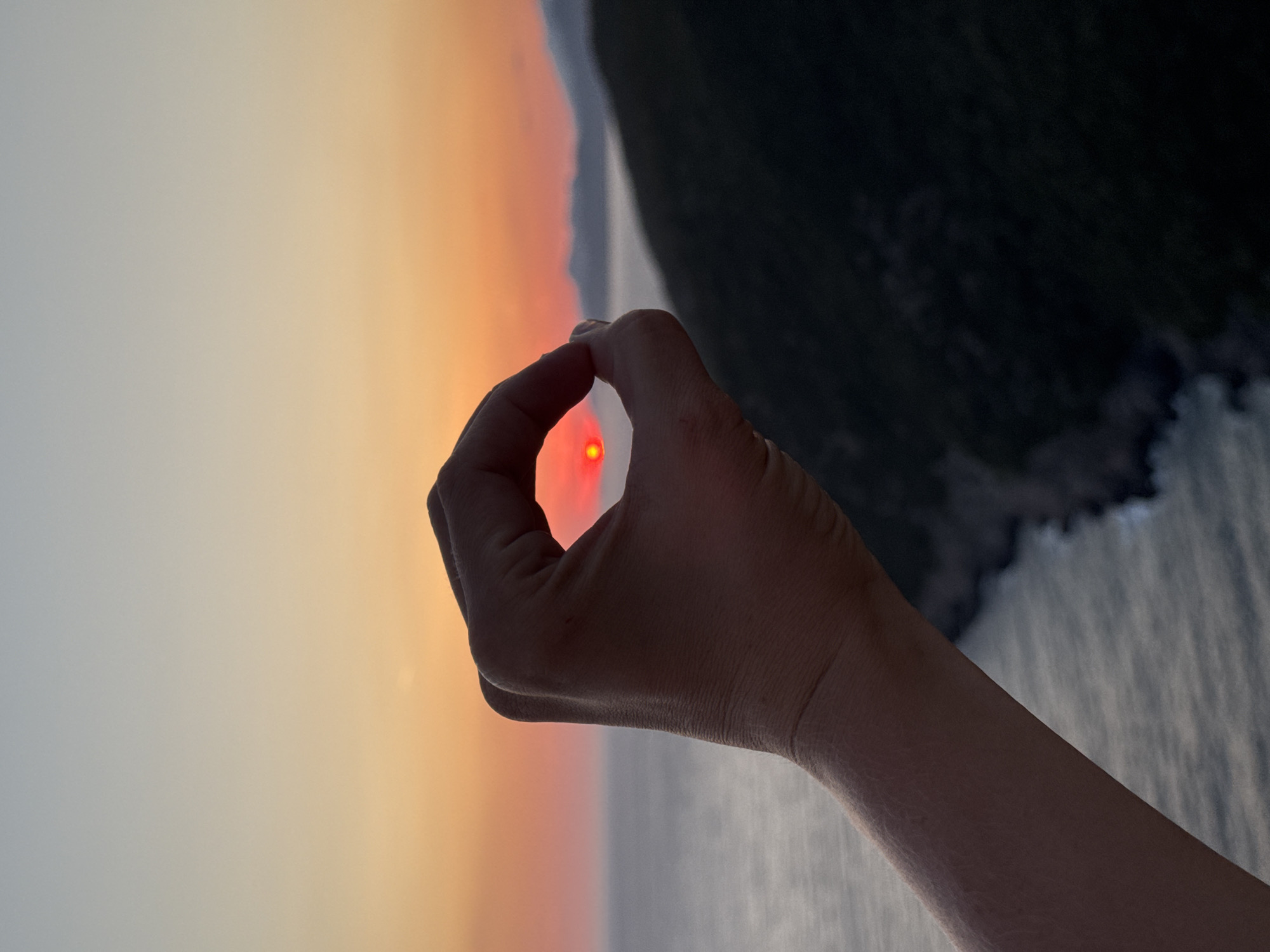A hand is held up in the foreground of the image, forming a circle with the thumb and index finger. Through this opening, the sun is visible, appearing as a small, bright red orb. The sun is low on the horizon, suggesting it is either sunrise or sunset. The background features a calm body of water, likely the Adriatic Sea, and a dark, hilly coastline. The sky displays a gradient of colors, transitioning from a pale blue at the top to orange and red near the horizon. The location is the coast near the city of Zagora, Montenegro, a region known for its picturesque coastal landscapes and dramatic sunsets. The overall impression is one of capturing a moment of natural beauty through a creative framing technique.