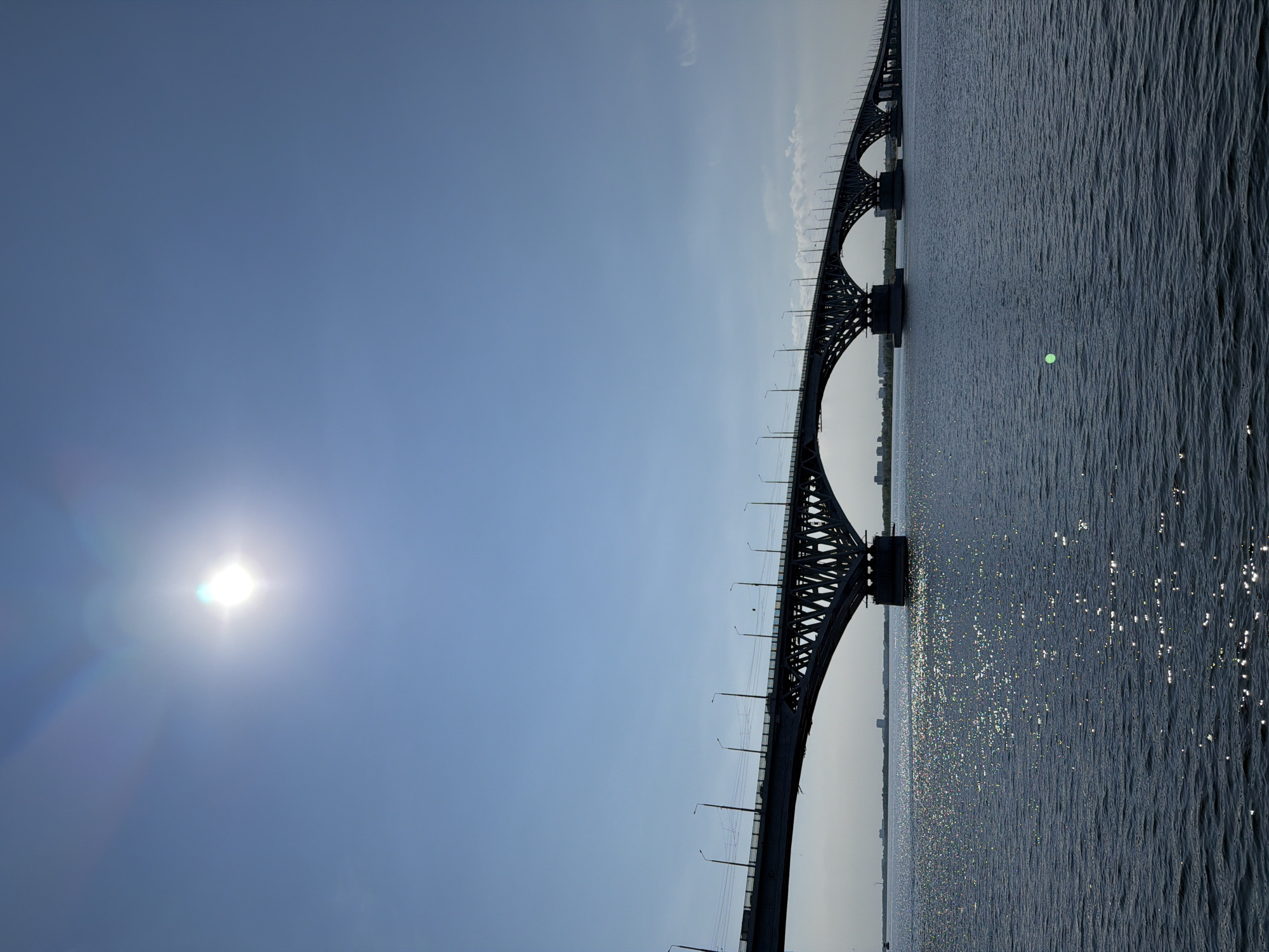 The image shows a large arch bridge spanning a wide body of water. The bridge is dark and silhouetted against a bright blue sky. The sun is visible in the upper left portion of the sky, with lens flare effects. The water is dark blue and reflects the sunlight in sparkling patches. In the background, along the horizon, is a city skyline with buildings. The bridge appears to be the Saratov Bridge, which crosses the Volga River, connecting Saratov and Engels in Russia. There are no people or specific actions depicted. The overall scene is one of a grand bridge and a sunny day over a river.