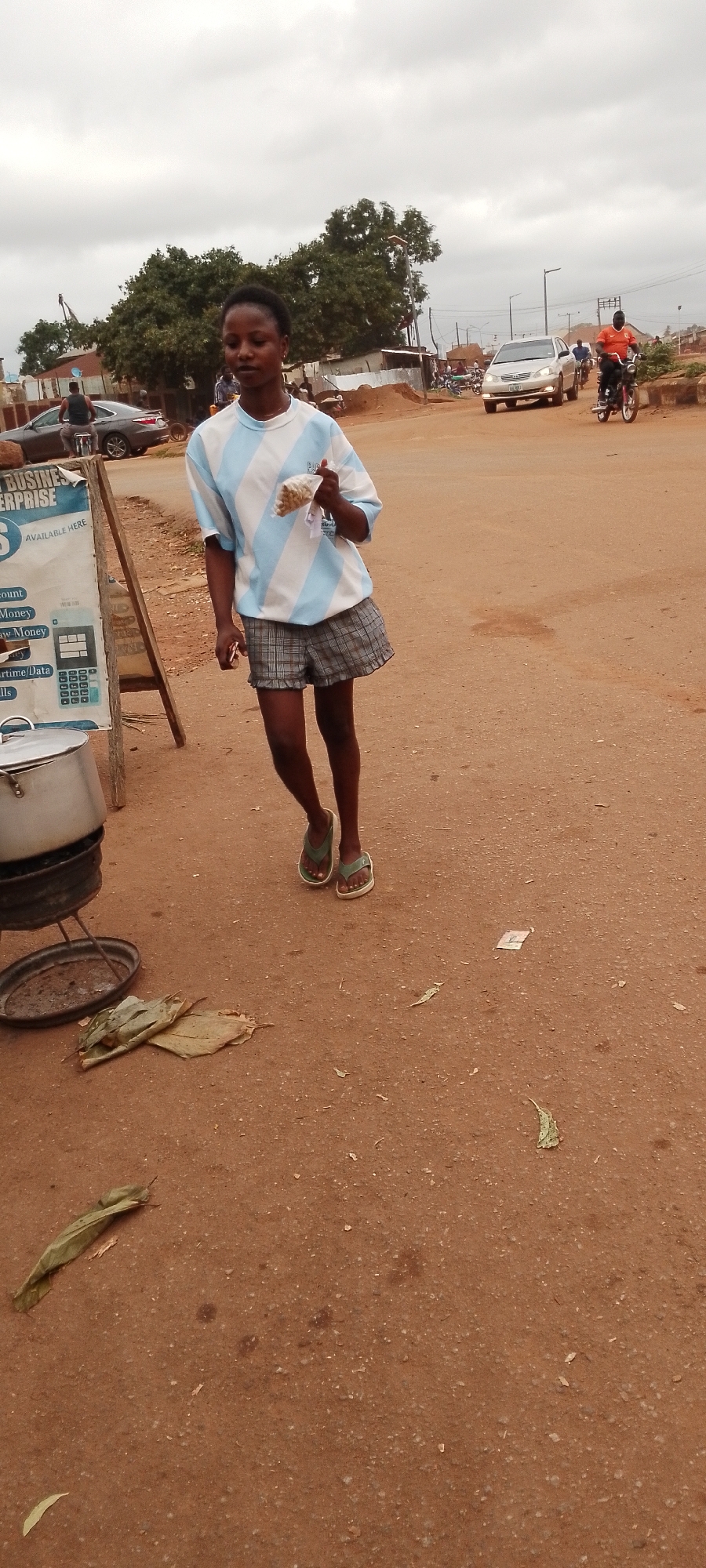 A young Black woman stands on a dirt road in Gboko, Nigeria. She wears a light blue and white striped t-shirt and plaid shorts. In her right hand, she holds a small plastic bag containing what appears to be food. Her left hand rests on her thigh. To her left, a wooden sign advertising business services and a large metal pot on a stand are visible. The background shows a street with vehicles, including cars and motorcycles, and buildings constructed of corrugated metal and brick. Trees and a cloudy sky complete the scene.