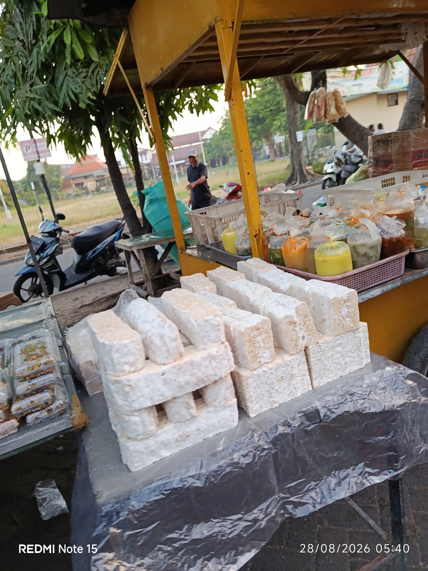 The image depicts an outdoor market stall, likely in Keboansikep, Indonesia, focusing on a selection of food items. In the foreground, large rectangular blocks of tempeh are neatly stacked, covered partially by a translucent plastic sheet. To the left, several clear plastic containers filled with what appear to be fried snacks or pastries are arranged. Behind these, a woven basket holds numerous plastic bags containing liquids of various colors, possibly drinks or sauces. In the background, a yellow-painted food cart or stall structure is visible, along with trees and a street scene. A person wearing a dark shirt is standing near the cart, and a blue scooter is parked on the side of the road. The overall impression is that of a vendor selling food at a street-side market.