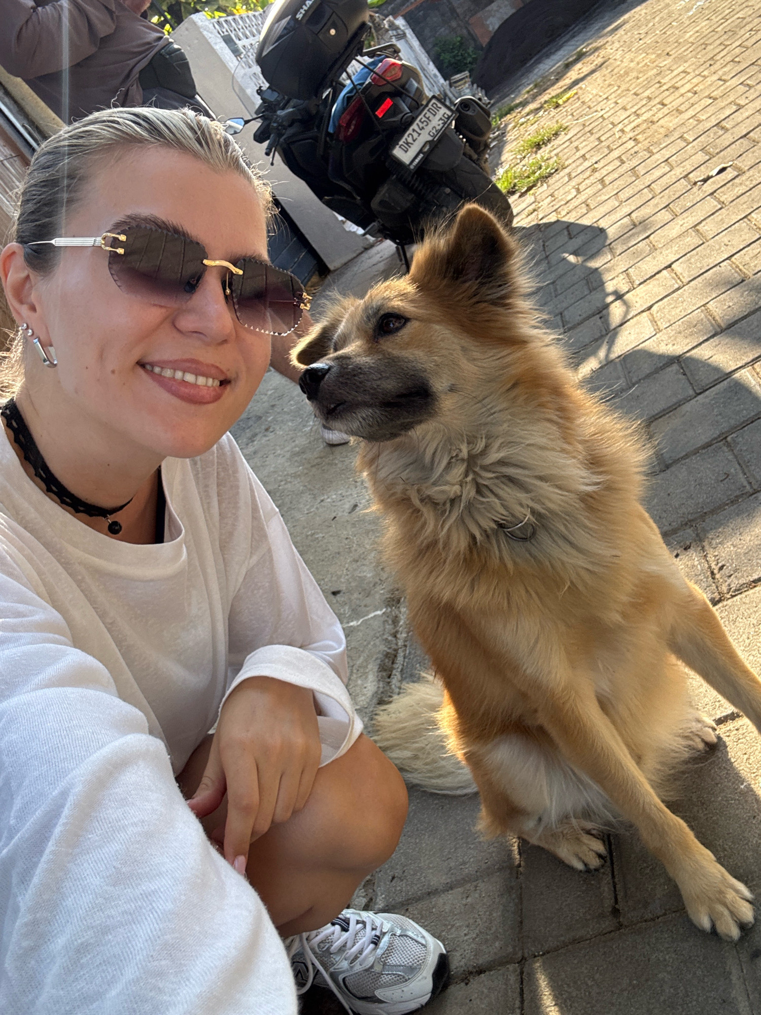 An outdoor, eye-level, medium shot captures a woman and a dog sitting on a paved walkway. The woman, in the foreground, faces the viewer and smiles. She has blonde hair pulled back, wears large, tinted sunglasses, a white long-sleeved shirt, and a black choker necklace with a black pendant. Her left hand rests on her knee, and her right hand is near her leg. Beside her, on the right, sits a medium-sized dog with light brown fur and a fluffy tail. The dog is looking to its left. Behind the woman and dog is a black motorcycle with a license plate that reads "DK2145FDR". The ground is paved with rectangular bricks, and sunlight casts shadows. In the upper left corner, a person wearing a dark shirt is partially visible, leaning against a structure.