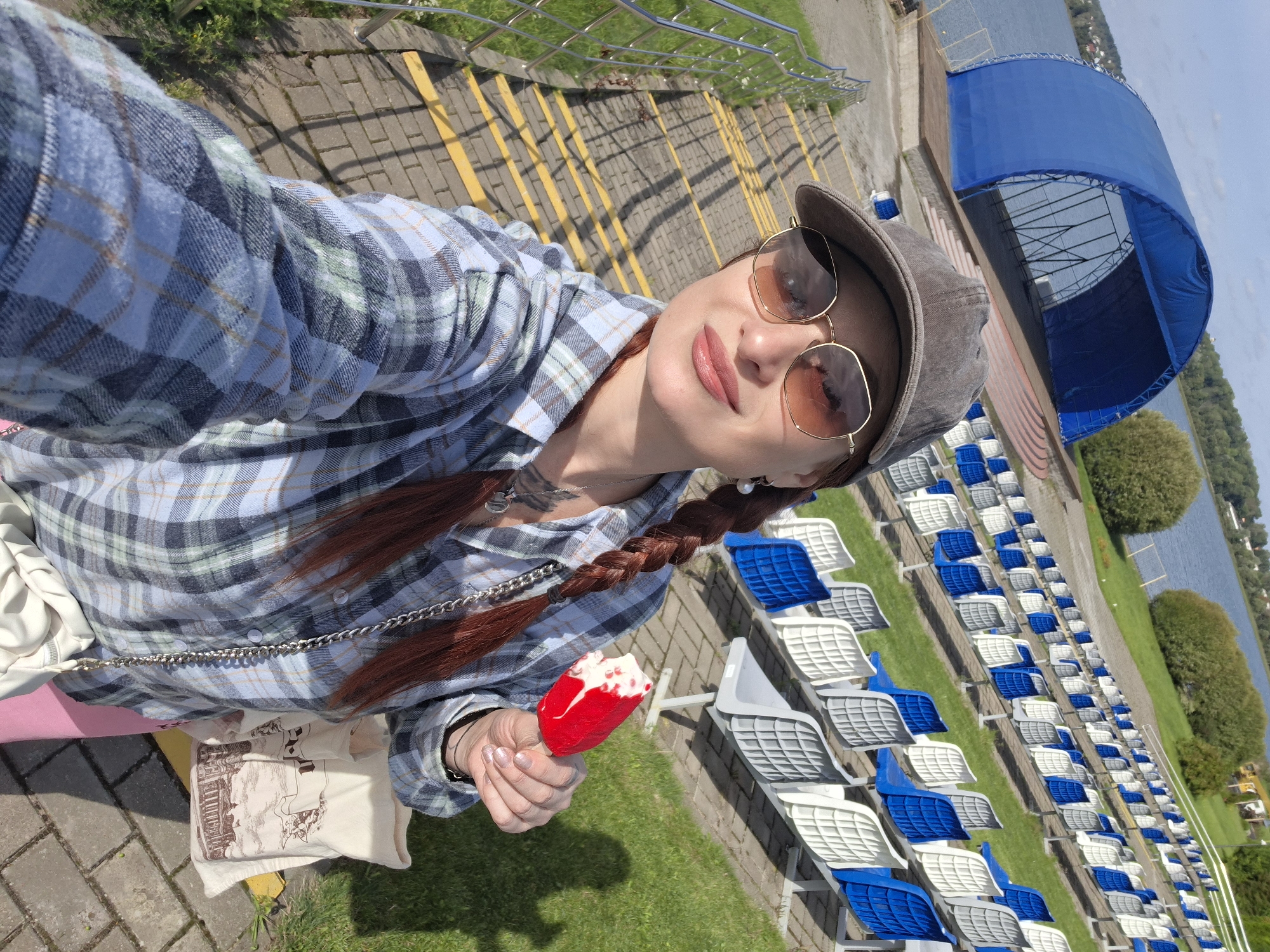 A woman with auburn hair in a braid is taking a selfie while holding a red and white popsicle. She is wearing a grey baseball cap, sunglasses, and a blue and green plaid shirt. She is standing on a paved walkway with yellow lines, next to a set of stairs with a metal railing. In the background, there is an amphitheater with blue and white seats facing a stage under a blue awning. A body of water and trees are visible beyond the amphitheater. The scene appears to be outdoors on a sunny day. This location is the Myadzyel City Beach Amphitheater in Myadzyel, Belarus.