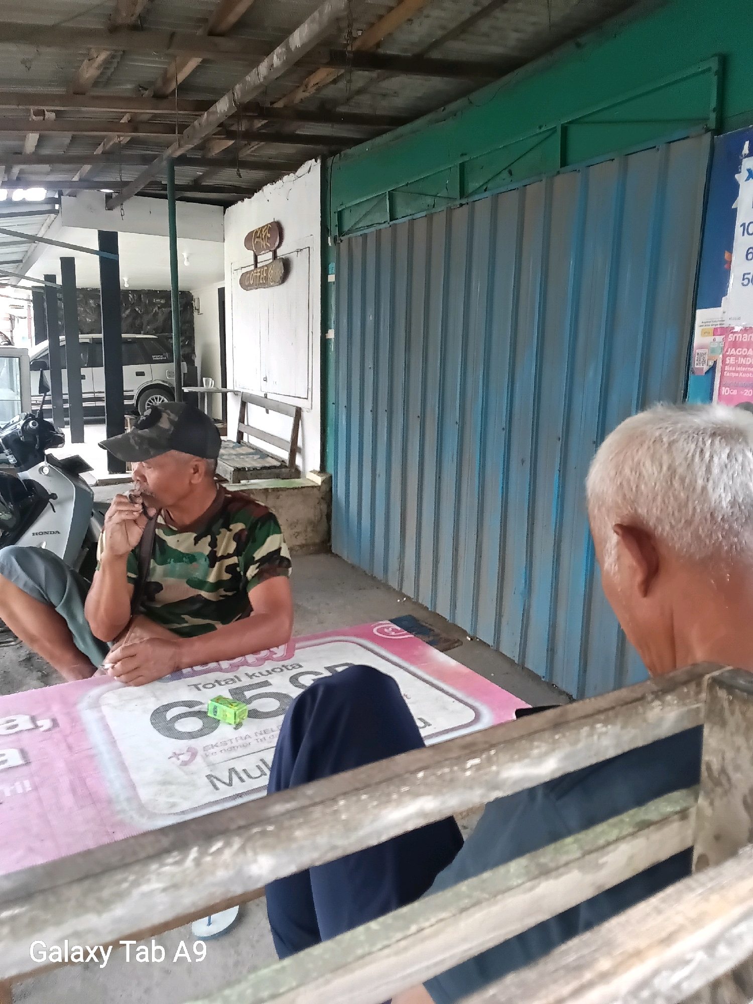 The image depicts an outdoor seating area, likely a roadside stall or eatery, in Wonoanti, Indonesia. Two men are present. In the foreground, an older man wearing a camouflage t-shirt and a black baseball cap is seated, smoking. He is holding a cigarette and appears to be looking downwards. To his right, another older man with white hair is visible from the back, facing away from the camera. He is seated and appears to be looking towards the left of the frame.
The environment suggests a casual, open-air setting. A table with a pink and white checkered surface is positioned between the men. On the table, a small green packet is visible. In the background, a light-colored vehicle, possibly a car or van, is parked. A motorcycle is also present to the left of the frame. The structure above them consists of a corrugated metal roof supported by wooden beams. Behind the men, a section of a building with a green corrugated metal facade is visible. There is also signage, including what appears to be a sign for "COFFEE" and another with text, possibly indicating prices or offers. The overall scene conveys a sense of everyday life in a provincial Indonesian town.