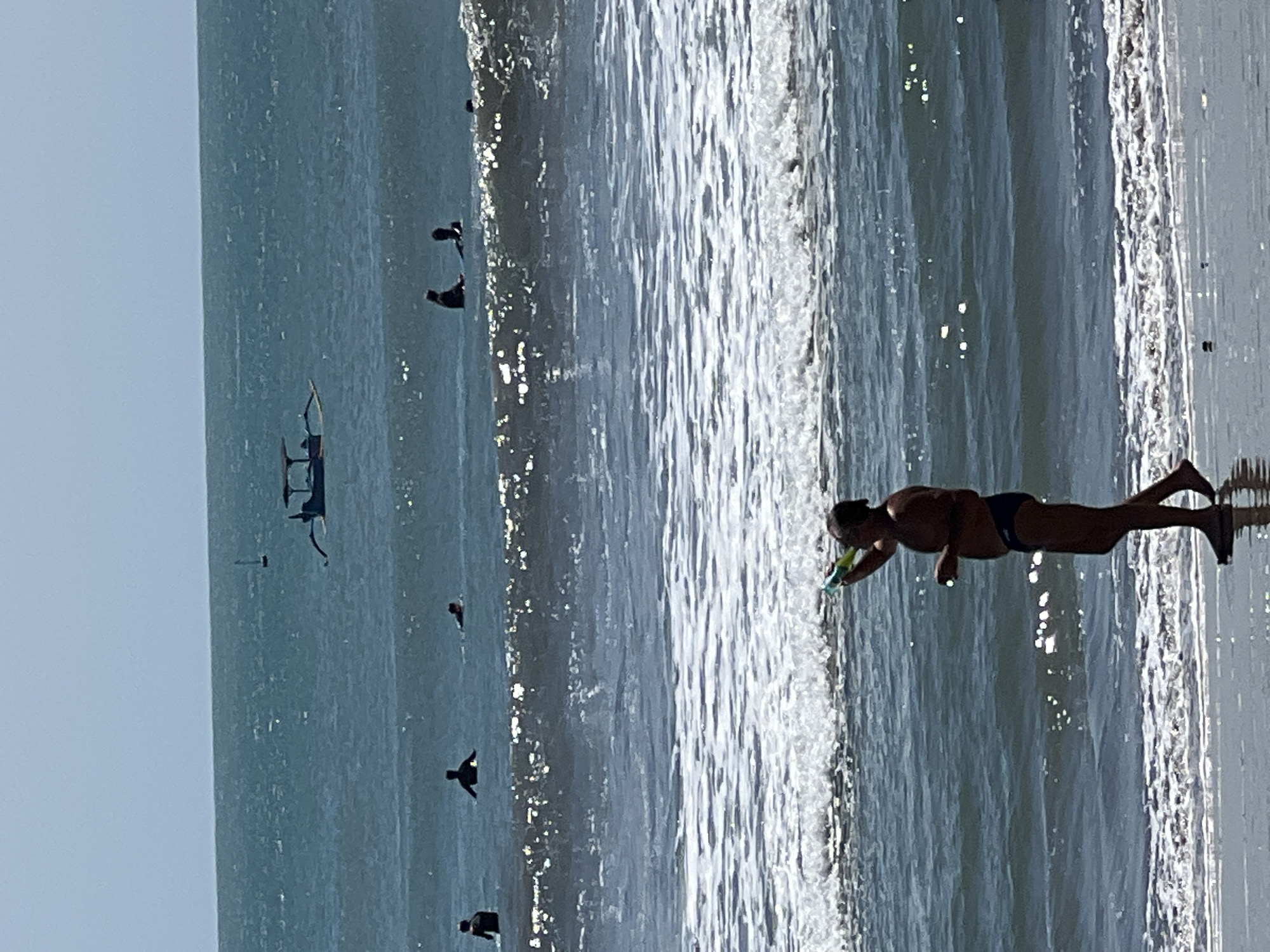 A man stands on the shore of a beach in Kuta, Indonesia, holding a colorful bubble gun and blowing bubbles. He is wearing dark swim trunks. In the background, several people are in the ocean, some appearing to be surfing. A small boat with a canopy is visible further out in the water, with a tiny white sail or flag. The water is a deep blue with moderate waves and sunlight glinting off the surface. The sky is a hazy light blue.