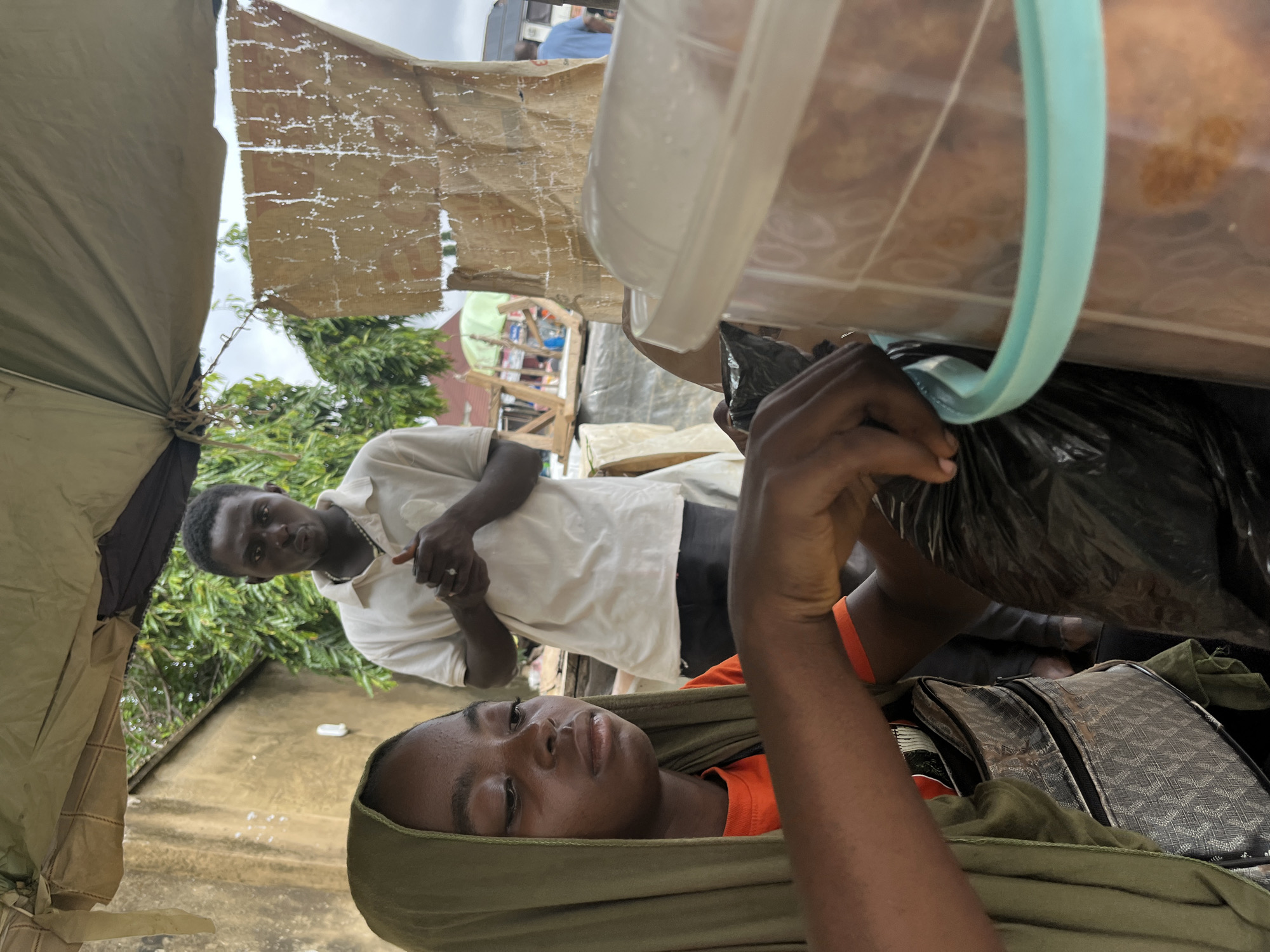The image depicts a street scene in Abuja, Nigeria. In the foreground, a young woman wearing a green headscarf and an orange shirt is seen from the chest up. She has her left arm extended and is actively engaged with a black plastic bag, appearing to be handling its contents. A portion of a patterned bag is visible beneath her arm, and a large, transparent plastic container with a light blue rim is situated to her right. Behind her, a young man in a white polo shirt stands with his hands clasped in front of him. He is looking directly at the camera with a serious expression. The background reveals a textured, light-colored wall, lush green trees, and what appears to be market stalls or shops, suggesting an outdoor commercial area. A glimpse of a bus and another person in a blue shirt can be seen further in the background, indicating the presence of ongoing activity in the vicinity. The scene is partially covered by a light-colored tarpaulin, suggesting it may be a stall or an improvised shelter from the elements.