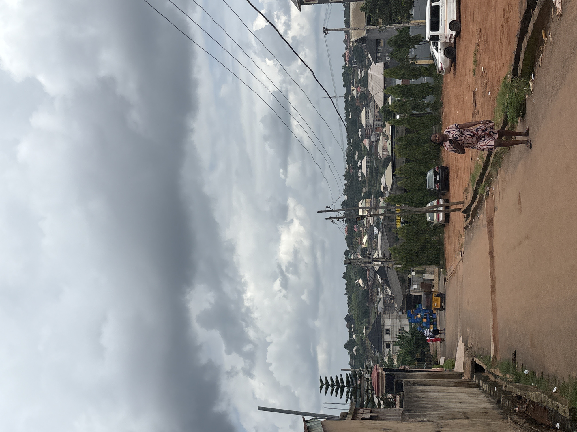 The media file depicts an outdoor scene in Nkpor, Nigeria. A woman in a patterned dress is walking along a dirt path. In the background, a dense urban landscape with houses and trees stretches across a hillside under a cloudy sky. Several vehicles, including a white van and other cars, are visible on the road. Power lines crisscross the sky, suggesting an inhabited area. The lighting suggests it is daytime, possibly late afternoon given the overcast conditions. The overall atmosphere appears to be that of a typical day in a bustling Nigerian town. There is some visible text on a yellow sign in the background, but it is illegible.