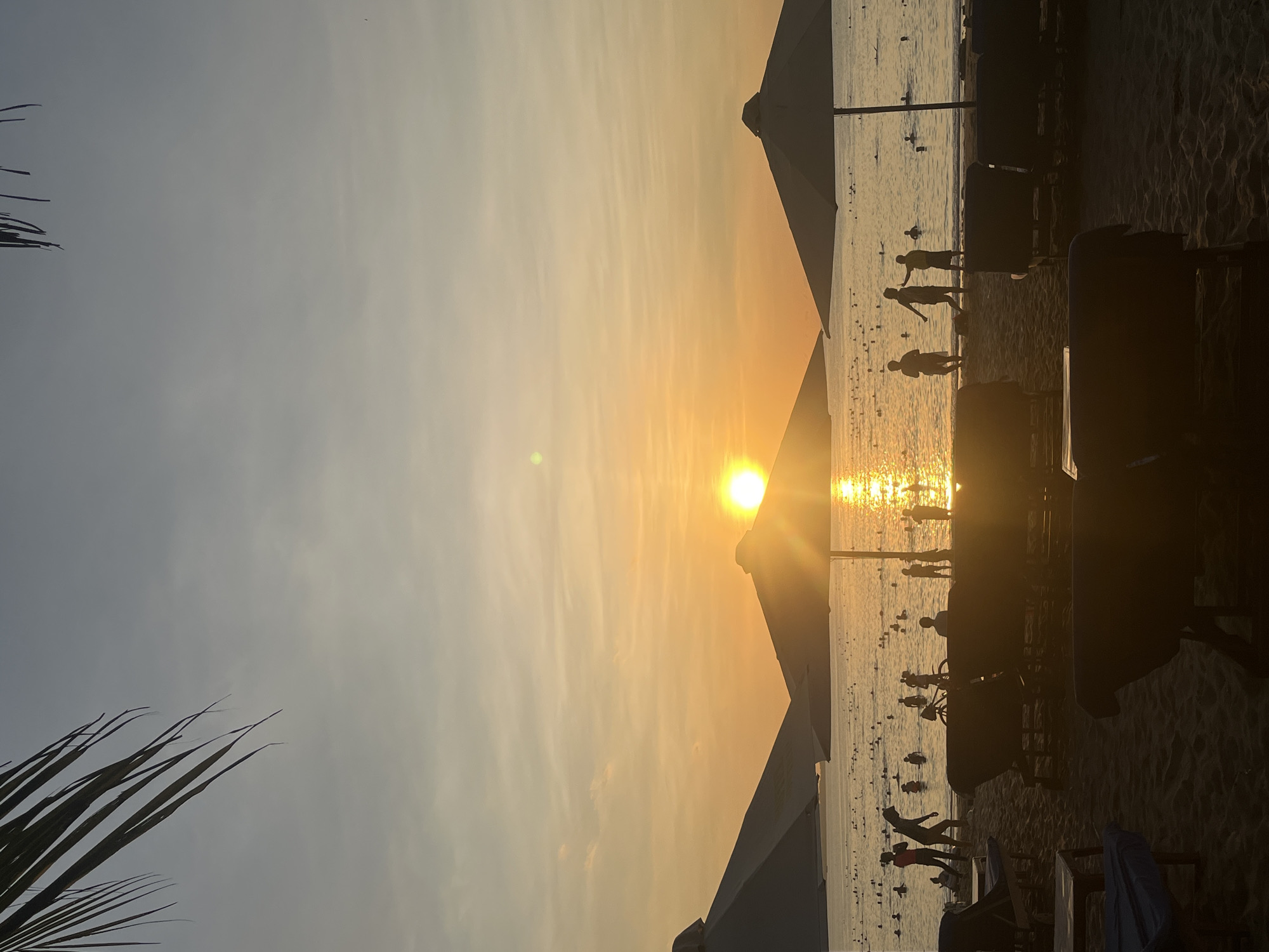 The image depicts a beach scene at sunset, potentially located in Bac Nha Trang Ward, Vietnam. The sun is positioned in the center, reflecting brightly on the water. There are people silhouetted along the shoreline and in the water. Beach umbrellas are visible, casting shadows. Beach chairs are also present in the foreground. Palm leaves appear on the left of the image.