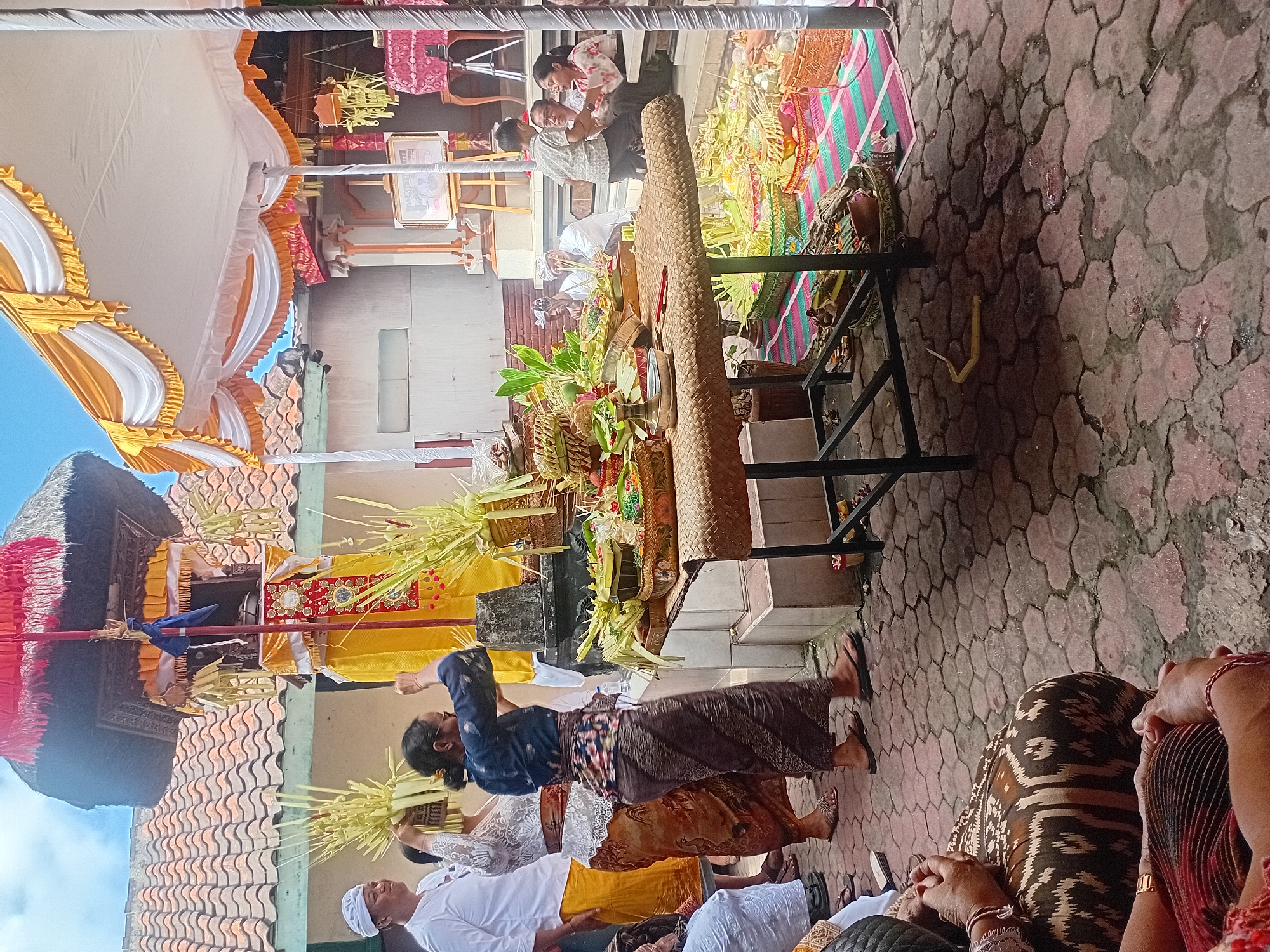 This image captures a scene of a religious offering ceremony, likely in Kuta, Indonesia. A group of people, dressed in traditional attire, are gathered around a low table laden with offerings. The offerings appear to be primarily composed of woven palm leaves, fruits, and other decorative items. Some individuals are actively participating in the ceremony, such as the woman in the foreground holding a woven basket of palm leaves aloft.
The environment features a traditional Indonesian architectural style, with tiled roofs visible in the background. A large white and yellow tent with decorative fabric is set up, suggesting an outdoor or semi-outdoor event. The ground is paved with hexagonal stones, some of which appear wet. In the background, a small altar or shrine is adorned with yellow and red fabric and decorative elements. Several individuals are seated or standing, observing the ceremony. A framed picture is displayed on an easel. The overall atmosphere suggests a communal and spiritual gathering.
