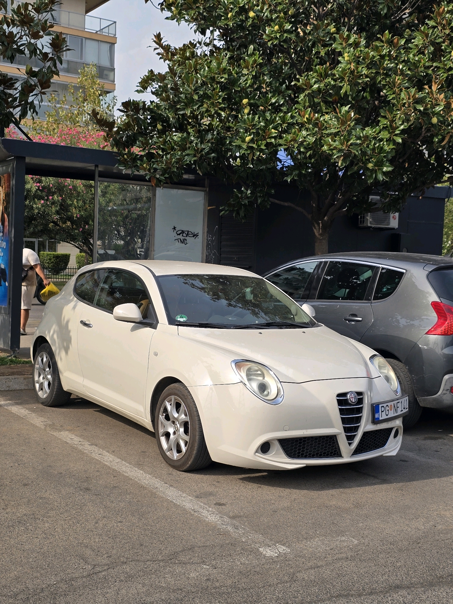 A cream-colored Alfa Romeo Mito is parked in a paved lot next to a grey Peugeot 207. The Alfa Romeo has a license plate that reads "PG NF 141". The car is facing towards the viewer, with its front end slightly angled to the right. The Peugeot is parked behind the Alfa Romeo and to its right, partially obscured. A person in a white t-shirt and beige shorts is visible walking behind the Alfa Romeo, carrying a yellow bag. The background includes trees, a building with balconies, and a dark structure with a window. The scene appears to be in a city.