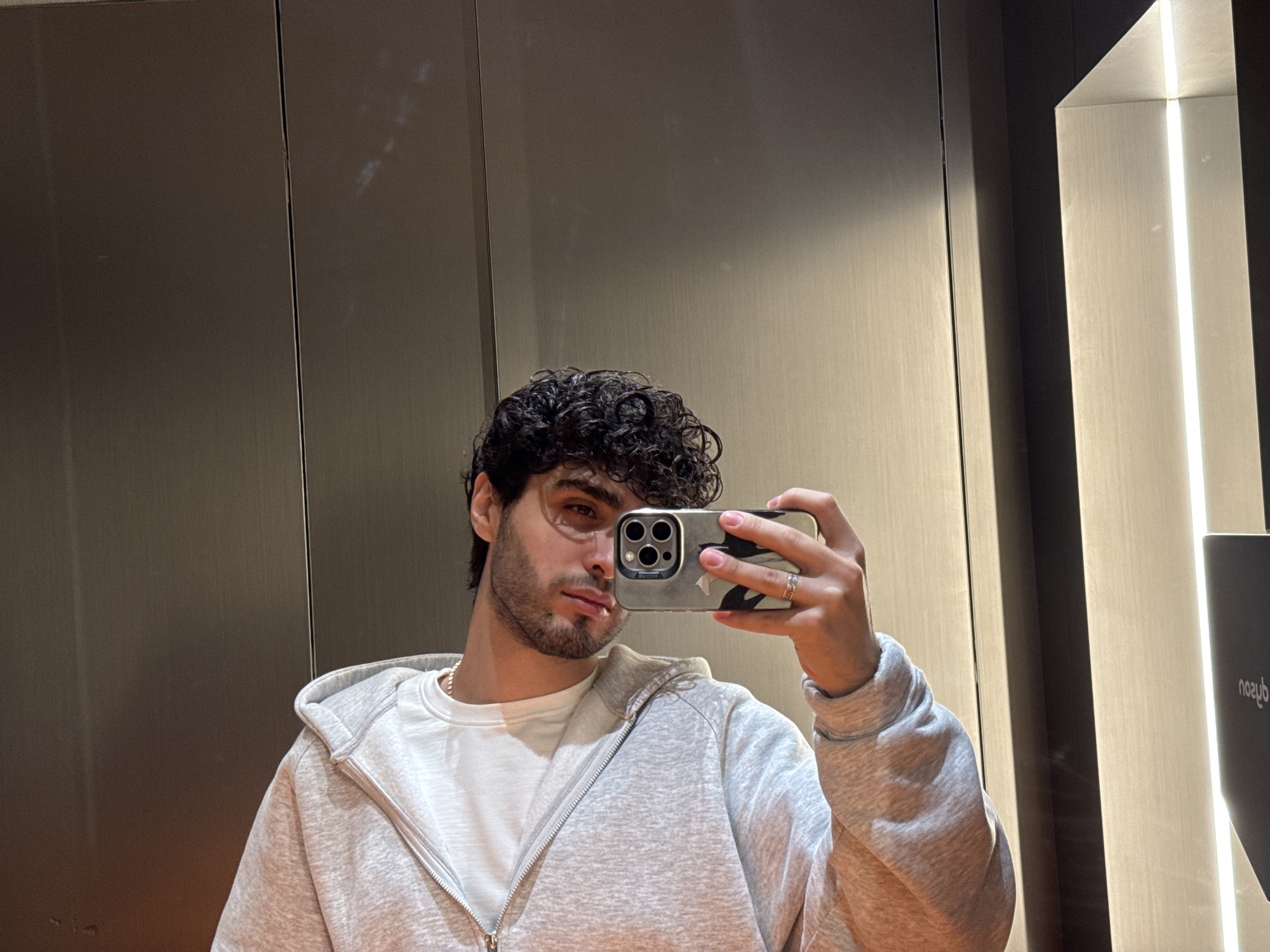 A young man with curly dark hair and a light beard is taking a selfie in what appears to be an elevator. He is wearing a white t-shirt under a light gray zip-up hoodie. He is holding a smartphone with a multi-lens camera in his right hand. He has a ring on his finger. The elevator has a modern interior with sleek, metallic walls and a vertical light panel. A screen is visible on the right side of the elevator. It is impossible to know whether this image was taken in the city of Yerevan, Armenia, without additional information.