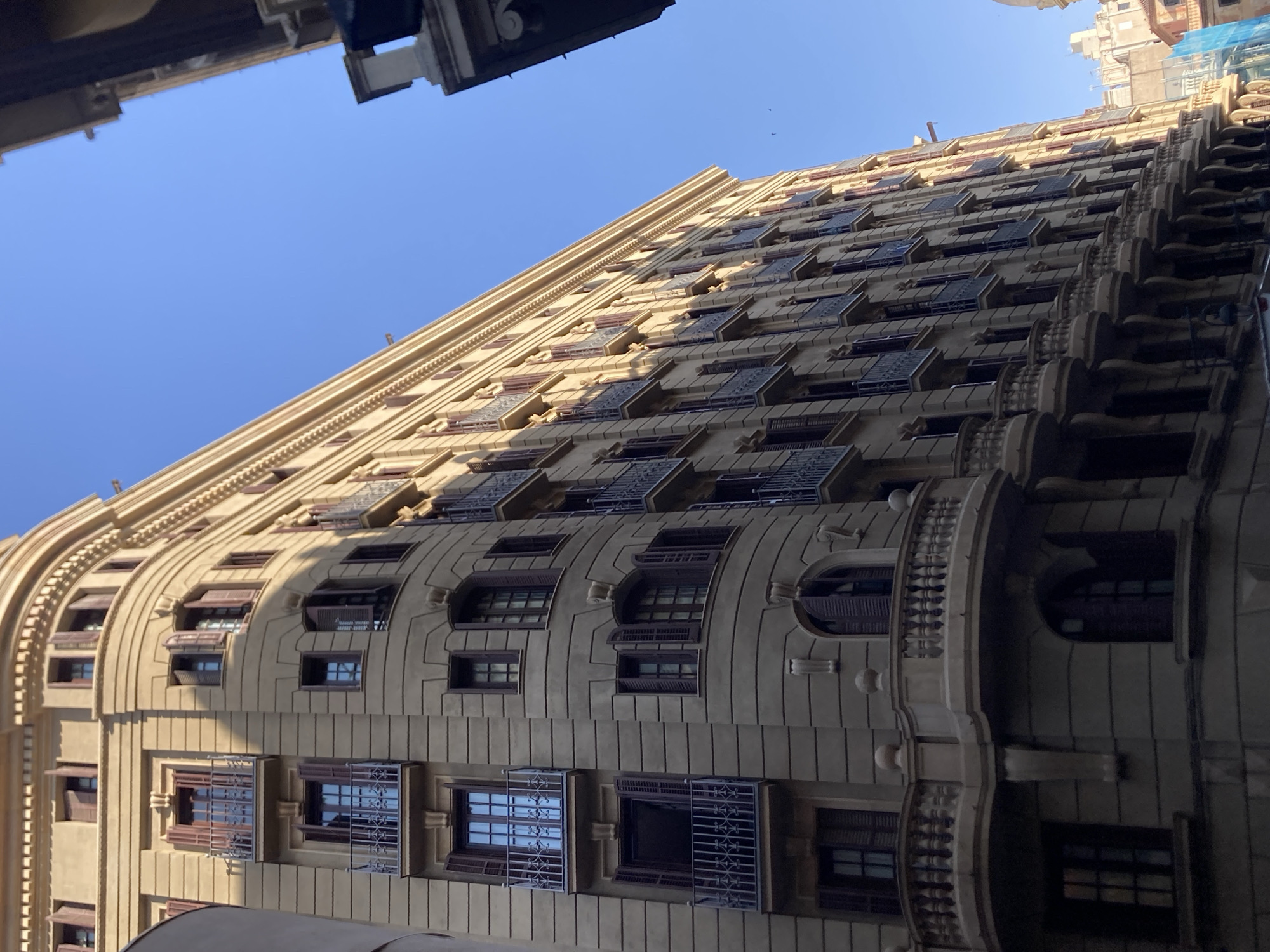 The image depicts a tall, ornate building with a curved facade, characteristic of Eixample architecture in Barcelona, Spain. The building is predominantly light brown, with multiple stories featuring numerous windows and balconies with wrought-iron railings. Many of the windows are open, revealing dark shutters. The building's facade is decorated with horizontal banding and decorative elements above some of the windows. The clear blue sky forms the backdrop. The perspective is from a low angle, looking upwards at the building, which is situated on a street where another building is partially visible on the right side, also appearing to be residential or commercial. The scene is devoid of any people.