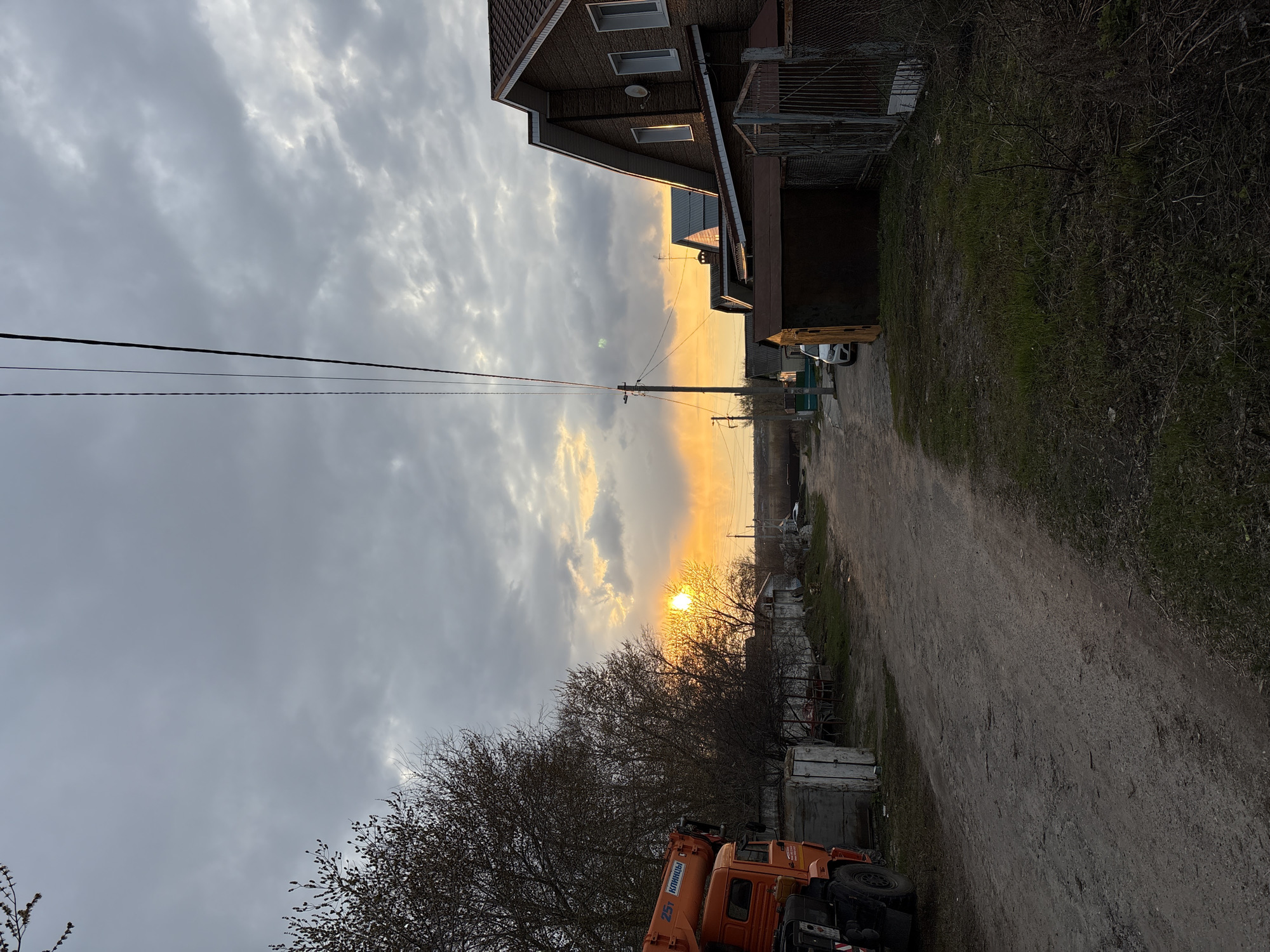 This media file captures an outdoor scene at either sunrise or sunset, characterized by a low sun casting an orange glow across a cloudy sky. The image appears to be taken along a dirt road or path, flanked by buildings and vegetation.
The sky is largely overcast with scattered, textured grey clouds. A bright, intense orange sun is visible through a break in the clouds, positioned roughly in the middle-right of the frame, illuminating the cloud edges with warm hues. Power lines stretch horizontally across the middle of the sky, supported by a utility pole visible on the right side of the road.
On the right side of the dirt road, several buildings are visible, including a prominent two-story house with dark brown siding and multiple rectangular windows under a gabled roof. Further down, other structures and fences can be seen. The road itself is unpaved, with visible tire tracks and sparse vegetation growing along its edges.
In the bottom left foreground, a large orange crane truck is partially visible. The text "КЛИНЦЫ" (KLINTSY) and "25Т" are clearly readable on its boom, indicating a Klintsy-made 25-ton crane. Further down the road on the right, a white car and a yellow heavy machinery vehicle (possibly an excavator) are discernible. Trees, some appearing bare or with early buds, are scattered throughout the scene, particularly a large cluster in the lower-middle and left. Grass and dry brush cover the ground in various areas. The overall impression is of a semi-rural or industrial fringe area in Ulyanovsk, Russia, during a peaceful late afternoon or early morning.